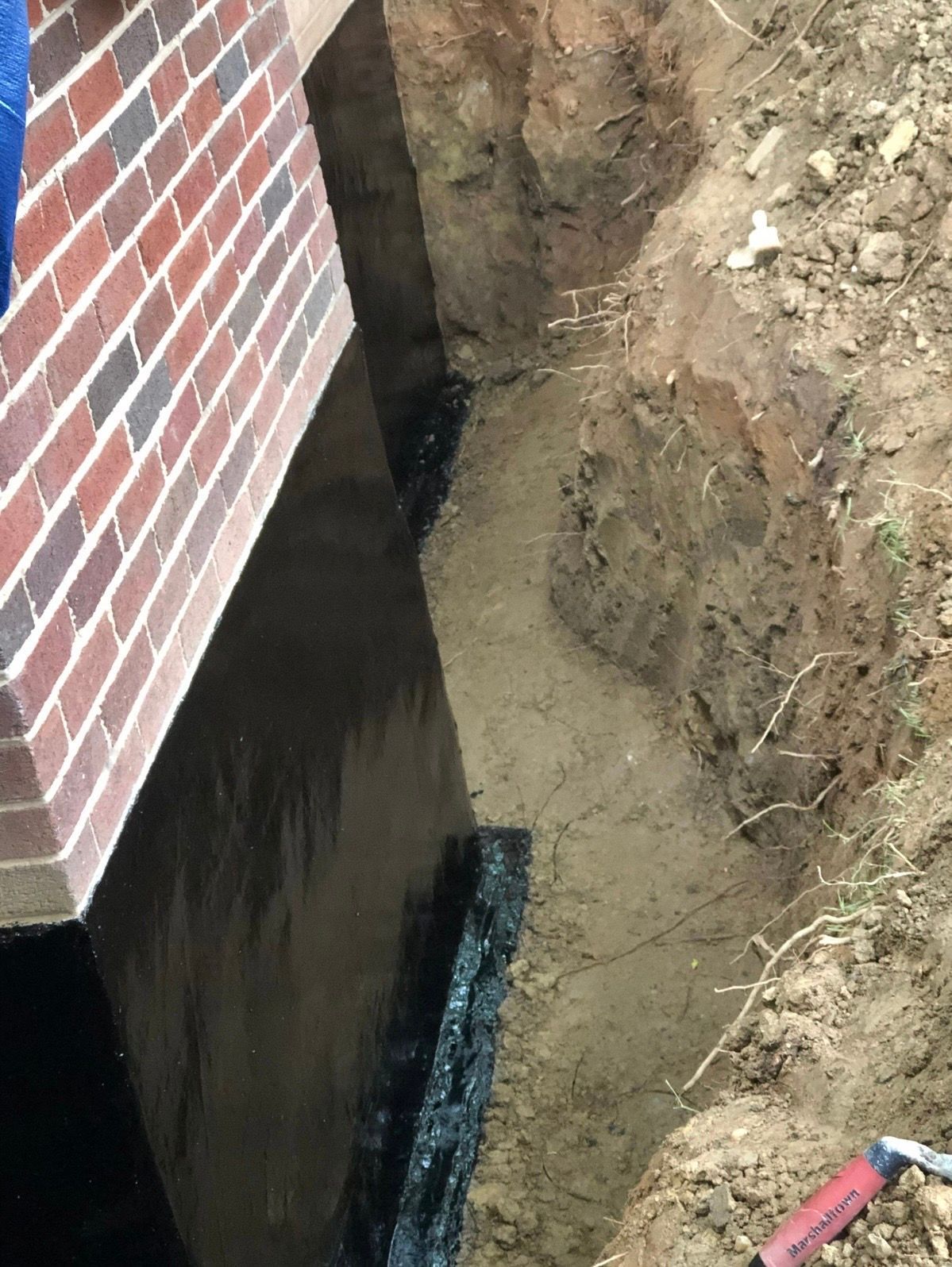 Narrow trench beside a brick house foundation, with exposed soil and a waterproofed black wall.