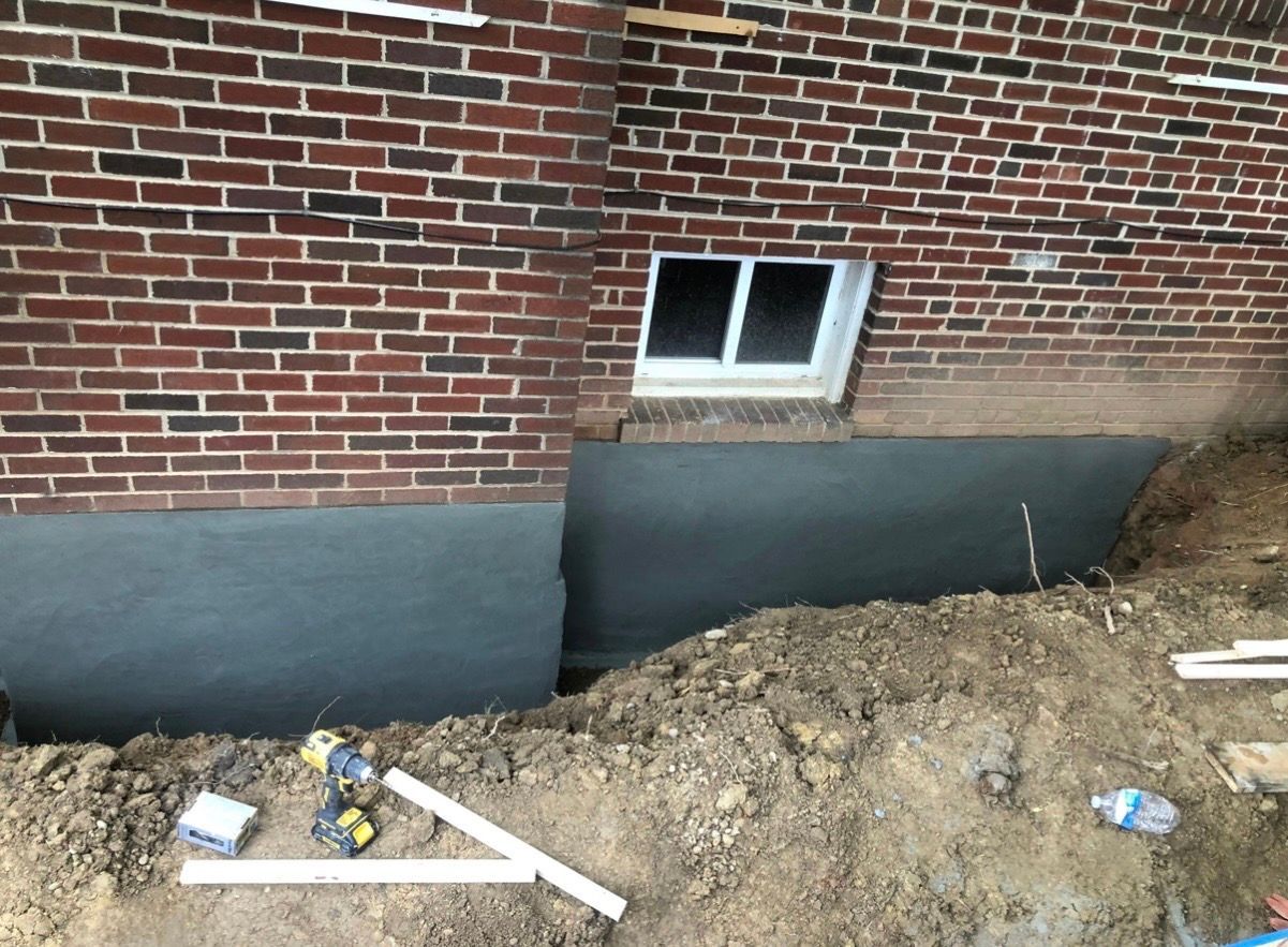 Brick house wall with a small basement window and fresh black foundation waterproofing above a dirt trench.