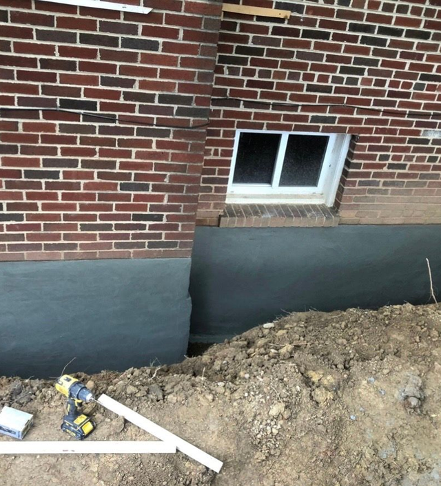Brick house wall with a basement window and fresh dirt trench along the foundation