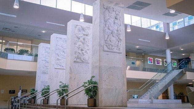 Bright atrium with tall carved pillars, indoor plants, skylights, and an escalator in a modern building.