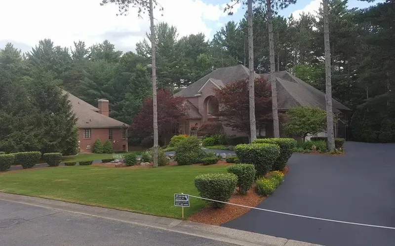 Two-story brick and dark shingle homes nestled in a wooded area. A paved driveway leads to the right. Manicured bushes line the yard.