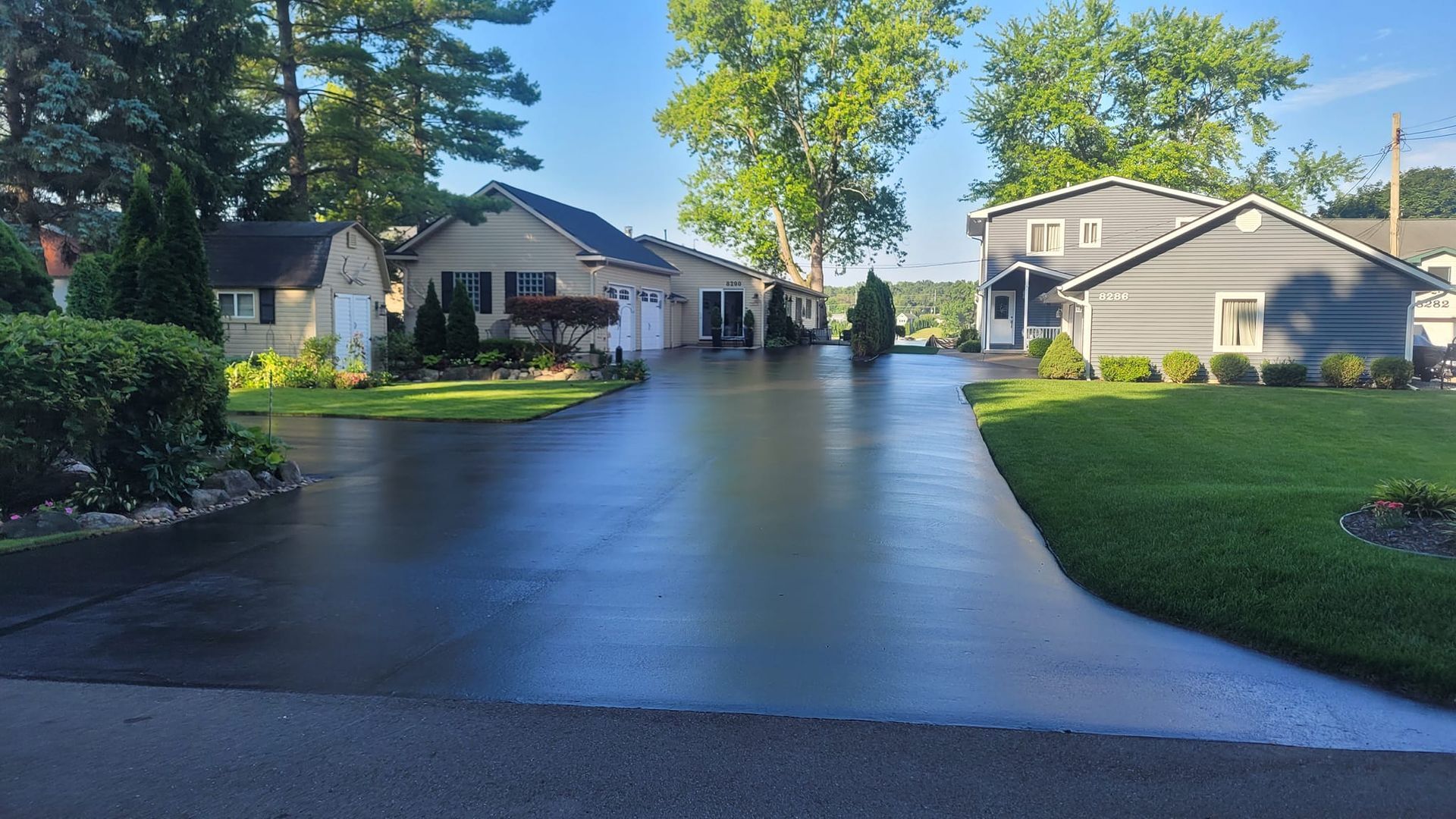 Paved driveway leading to multiple houses along a lake. Green grass and trees surround the homes under a blue sky.