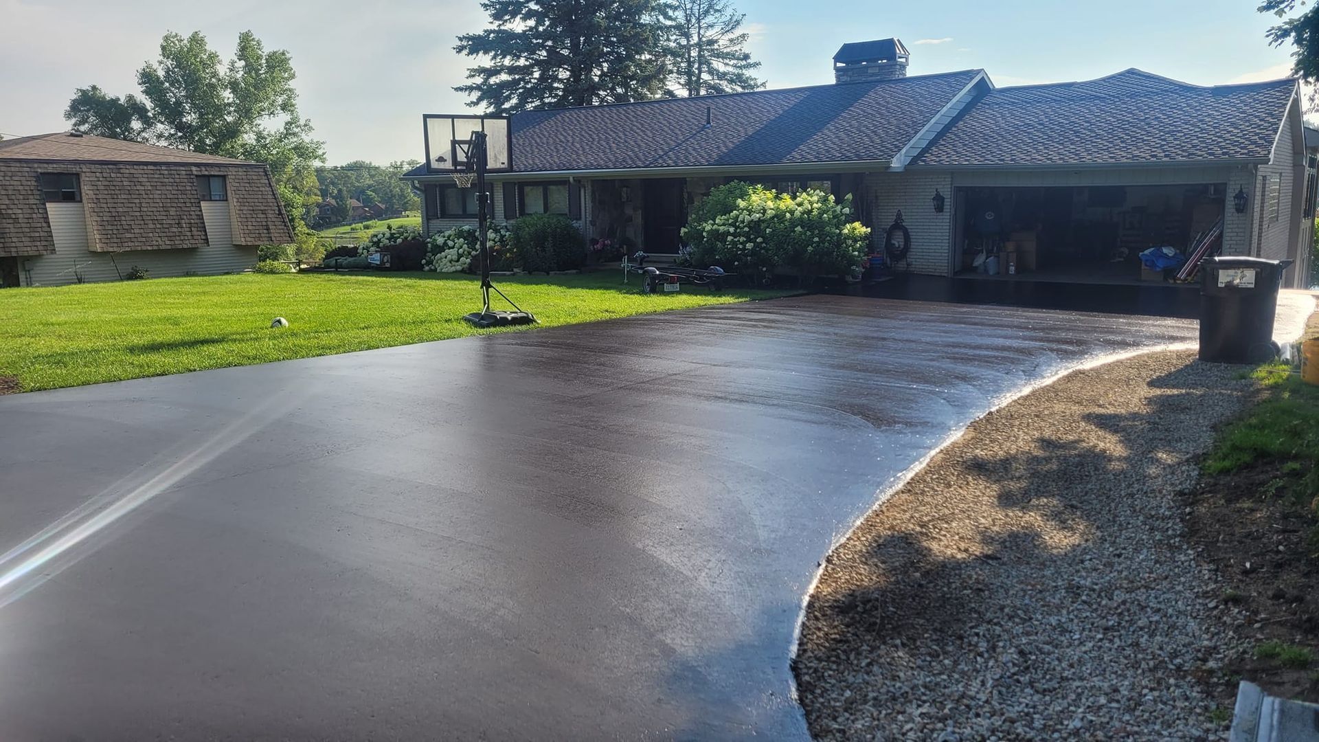 A wet, black driveway in front of a single-story house with a garage and basketball hoop. The sky is blue and a grassy area is on the left.
