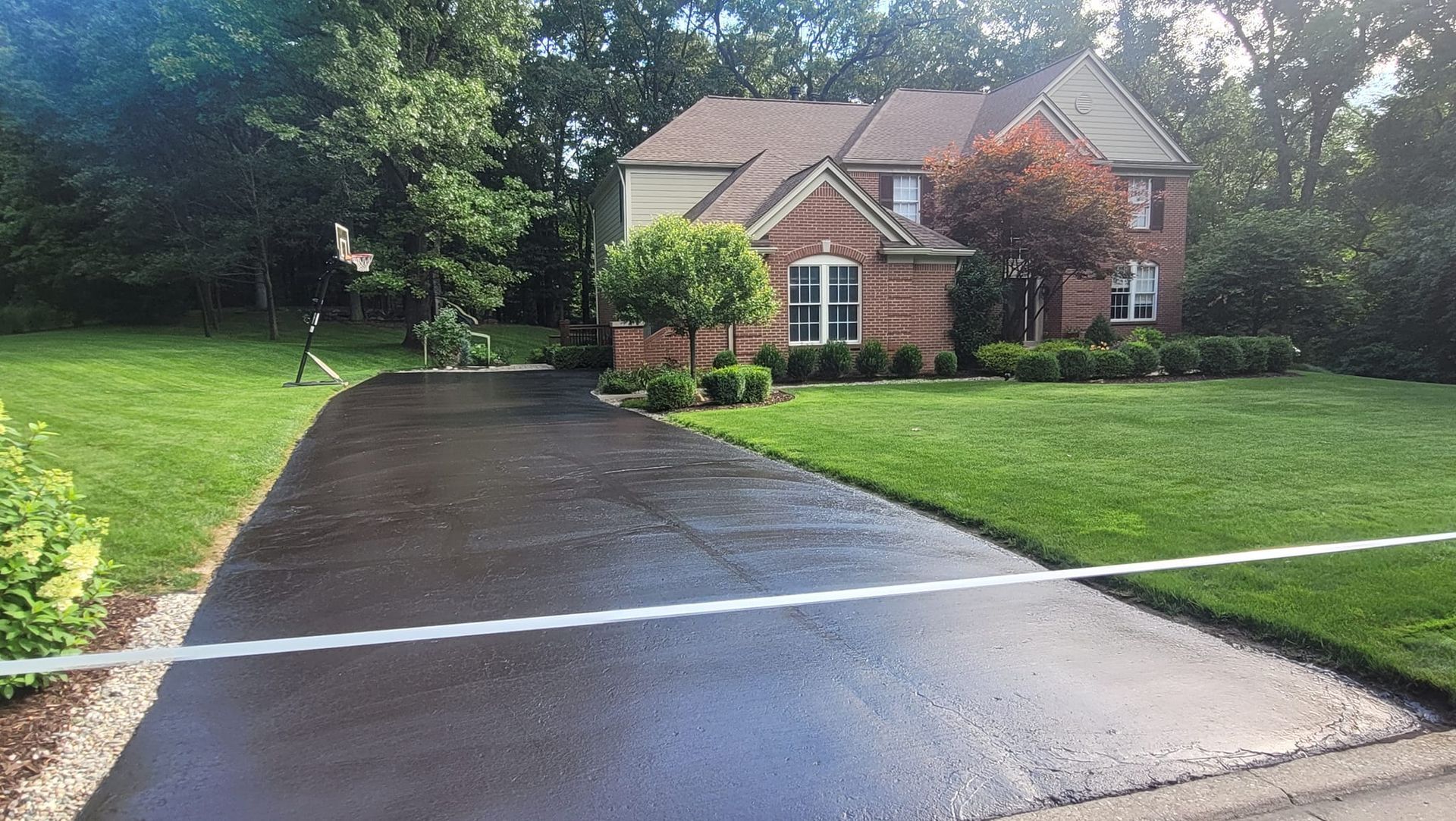 A newly sealed asphalt driveway leads to a two-story brick house with lush green lawn and trees.