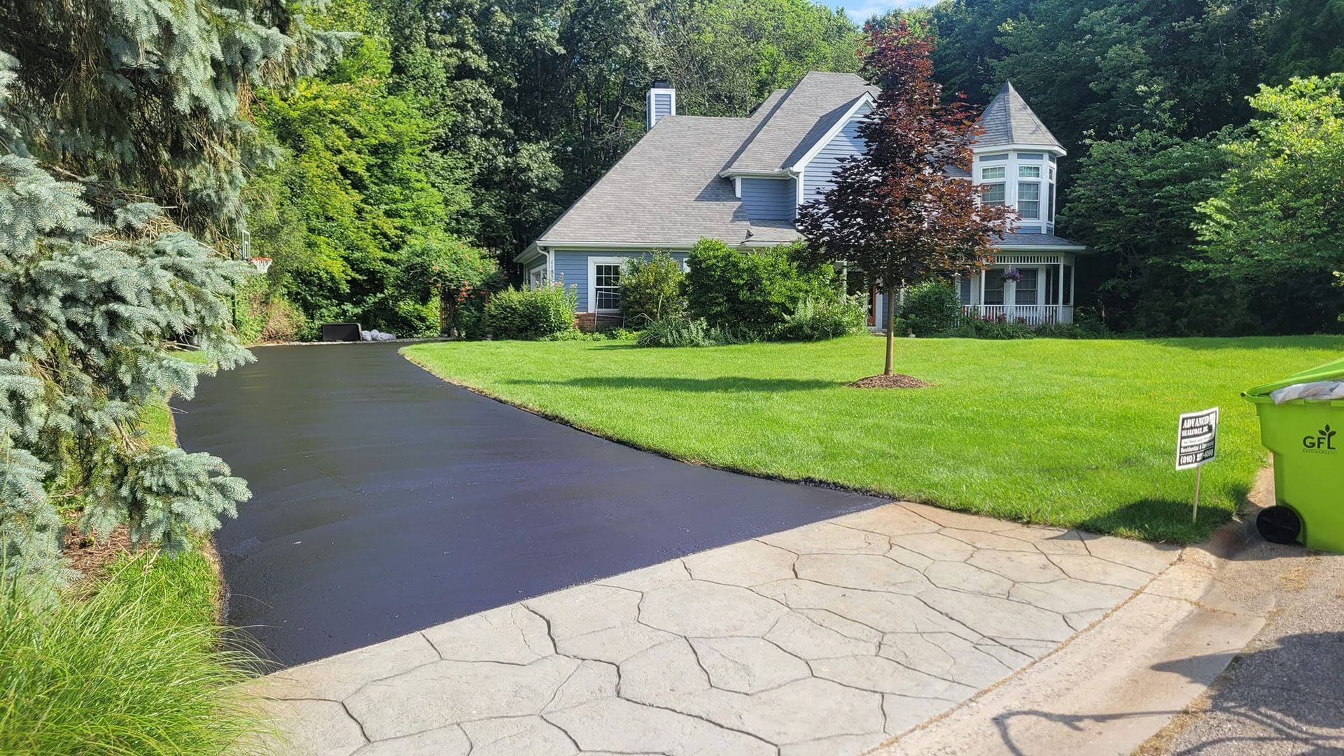 A blue house with a gray roof and a black asphalt driveway is surrounded by green grass and trees.