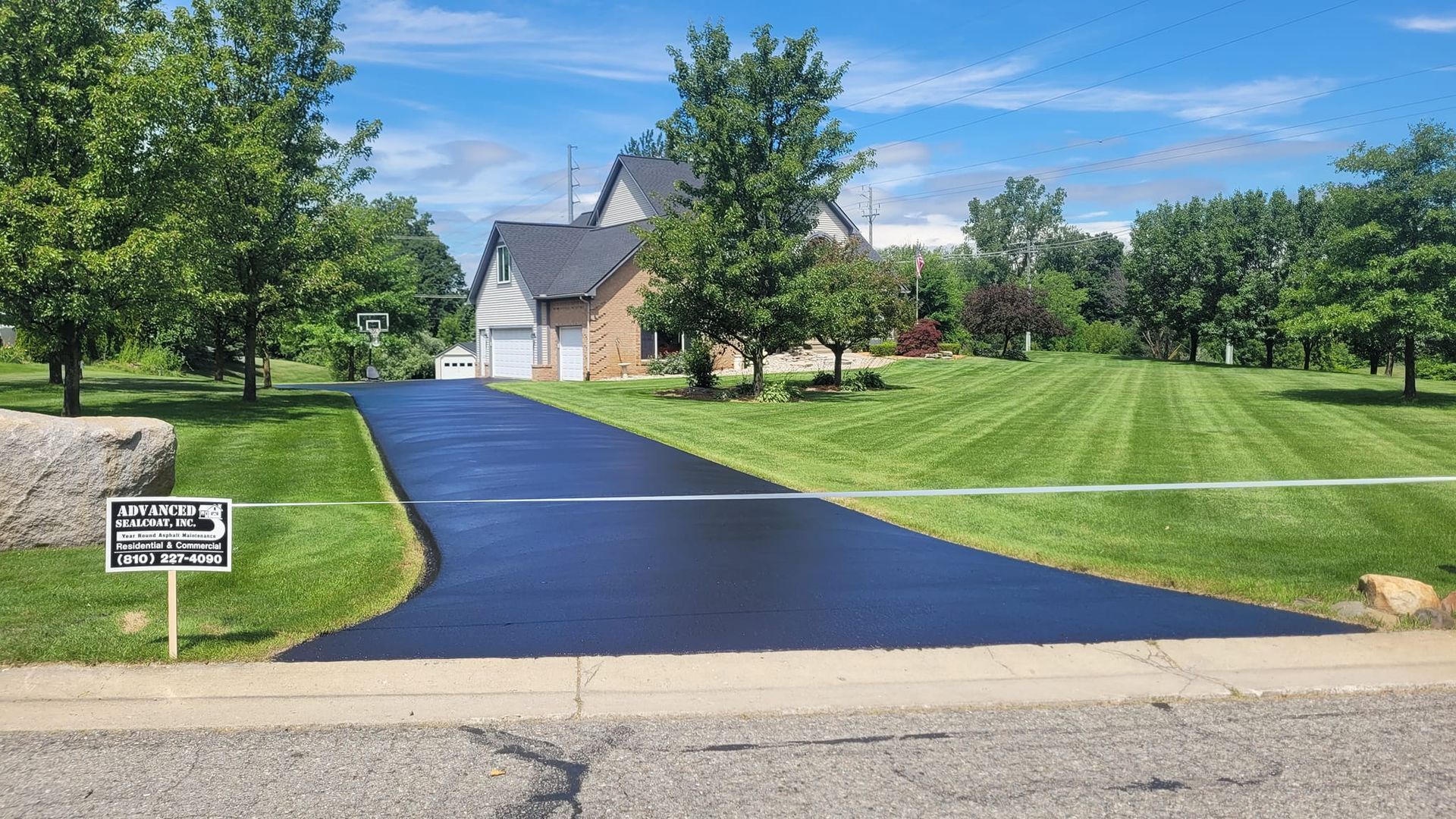 Newly paved asphalt driveway leading to a light brick and gray-roofed house, surrounded by green grass and trees.