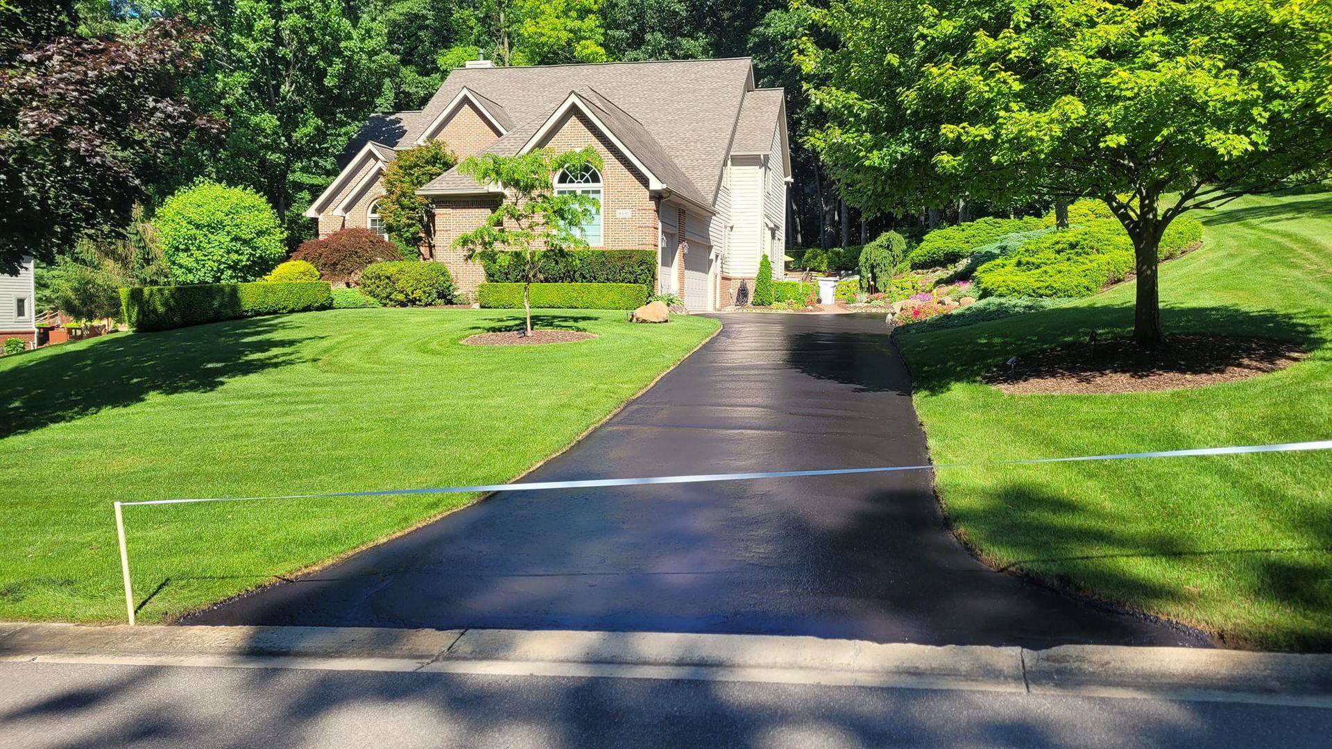 A newly sealed black asphalt driveway leading to a house with a brown roof, surrounded by green grass and trees.