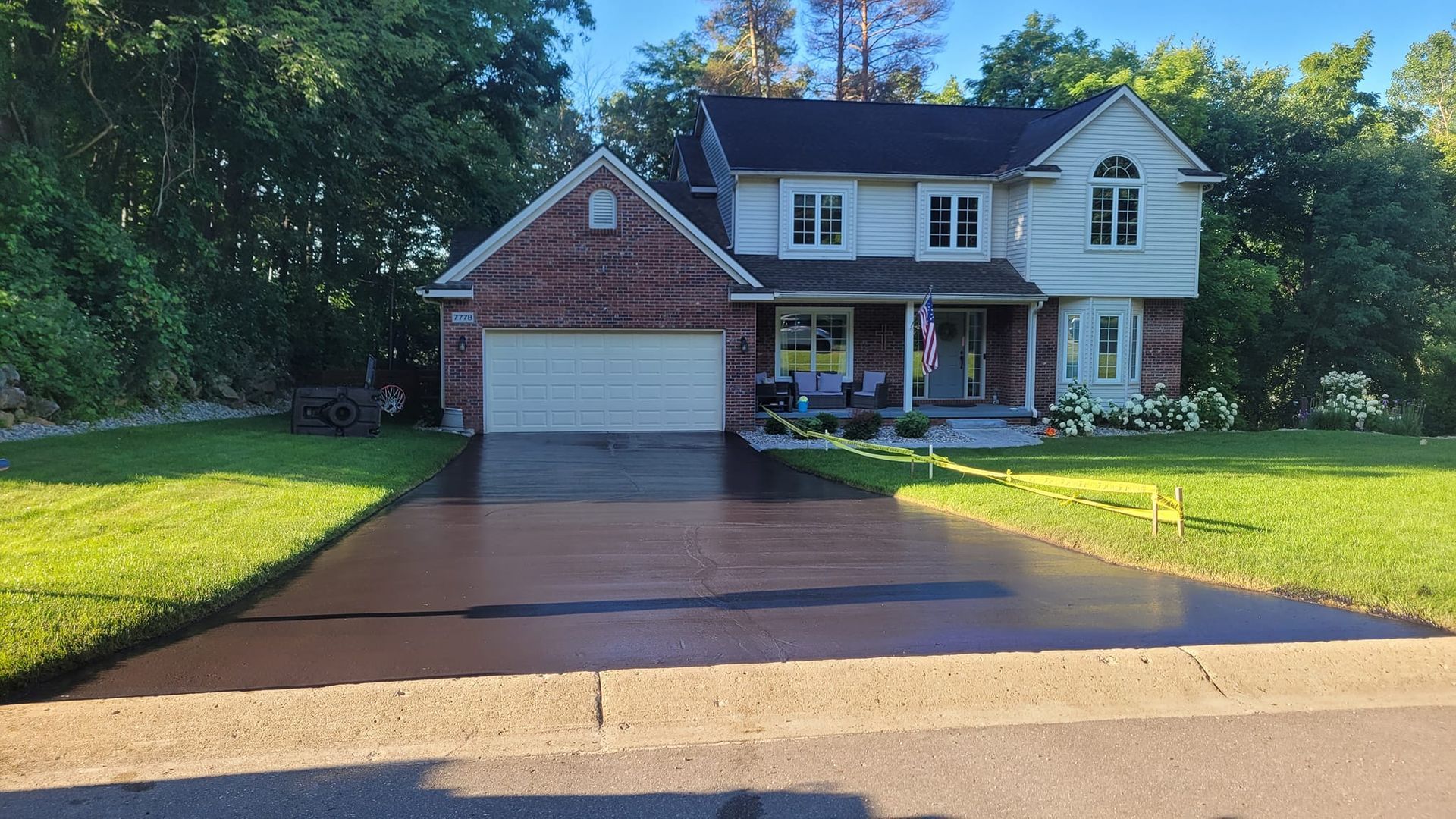 A two-story house with a freshly sealed black driveway, flanked by green lawns on a sunny day.