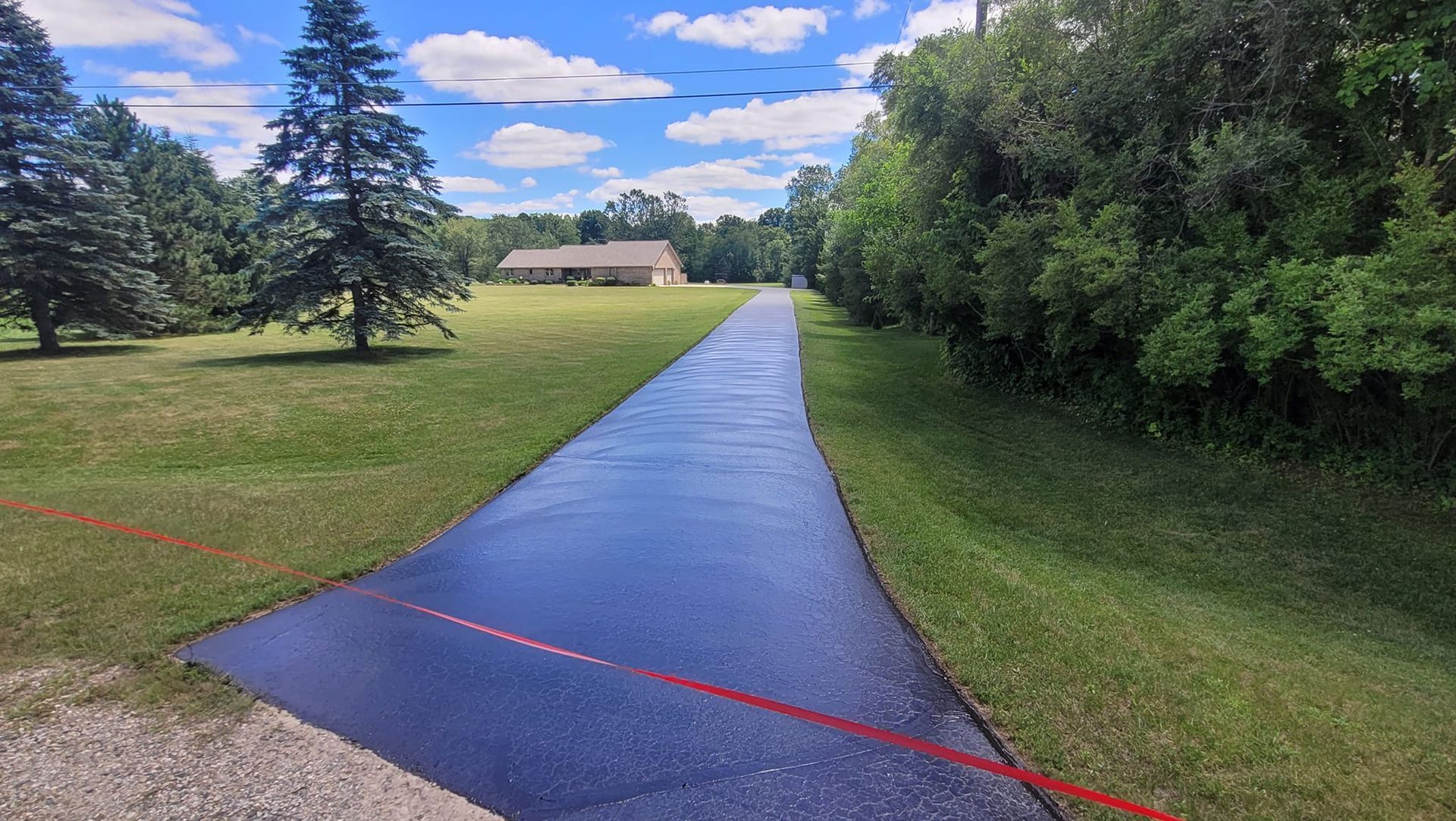 Long, black asphalt driveway leading towards a house, flanked by green grass and trees under a bright blue sky.