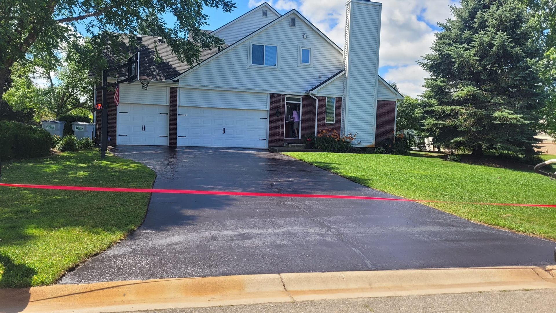A two-story white house with a dark driveway, green lawn, and a red line across the driveway. The open garage door reveals someone inside.