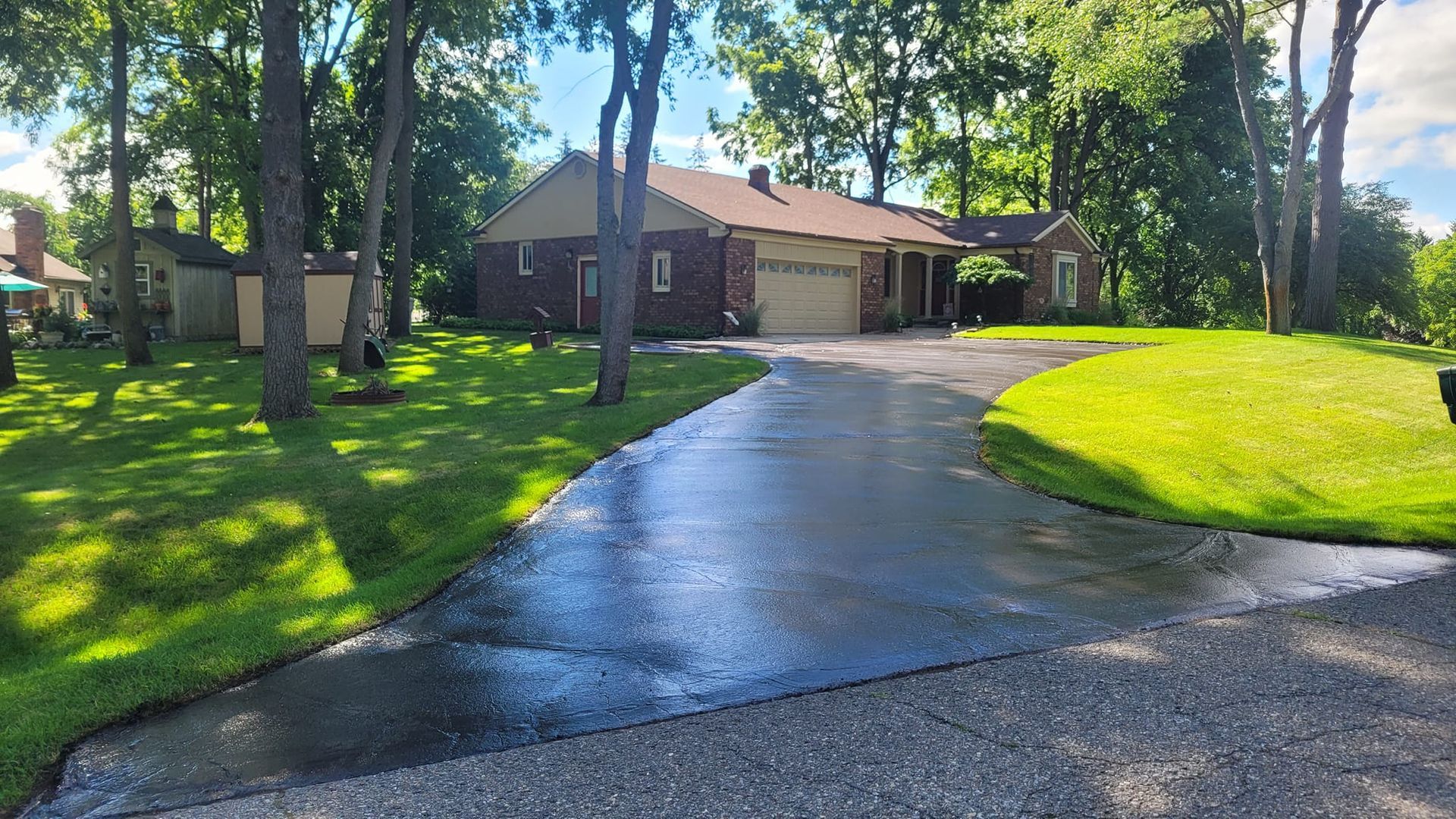 Asphalt driveway leading to a brick house with a two-car garage, surrounded by green grass and trees. The driveway appears recently sealed and wet.