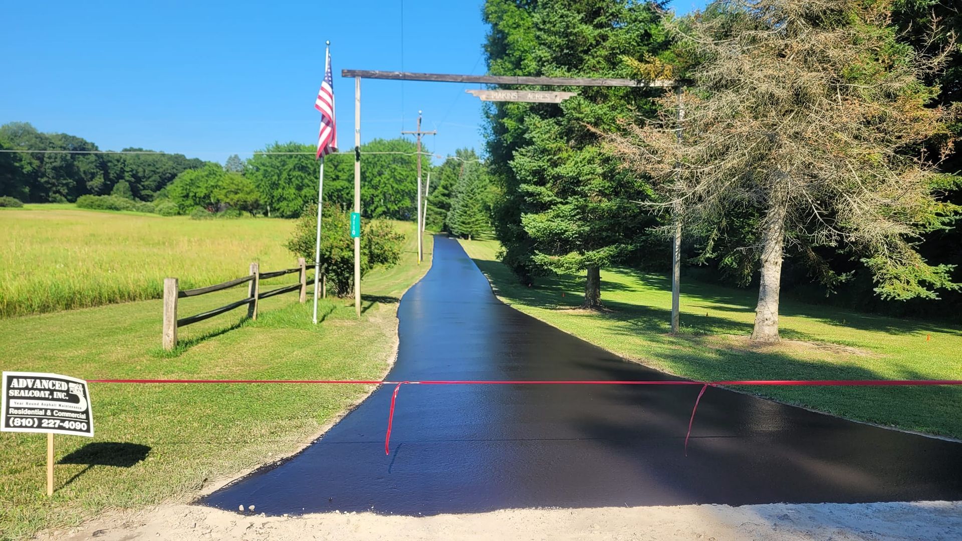 Newly paved asphalt driveway leading into a rural property, marked with a red line. An American flag and trees line the entrance.