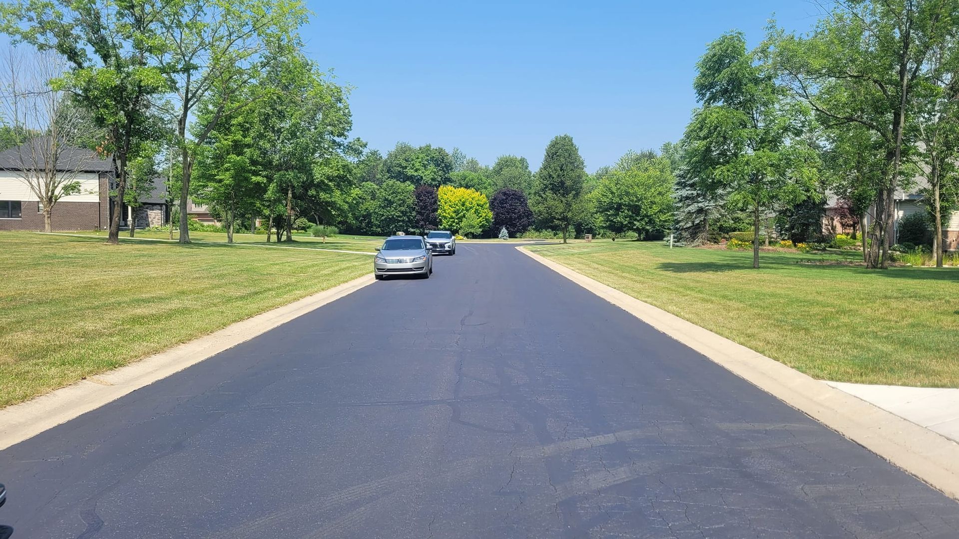 A paved residential street with two cars driving down the middle; lined with green grass, trees, and houses. Clear blue sky.