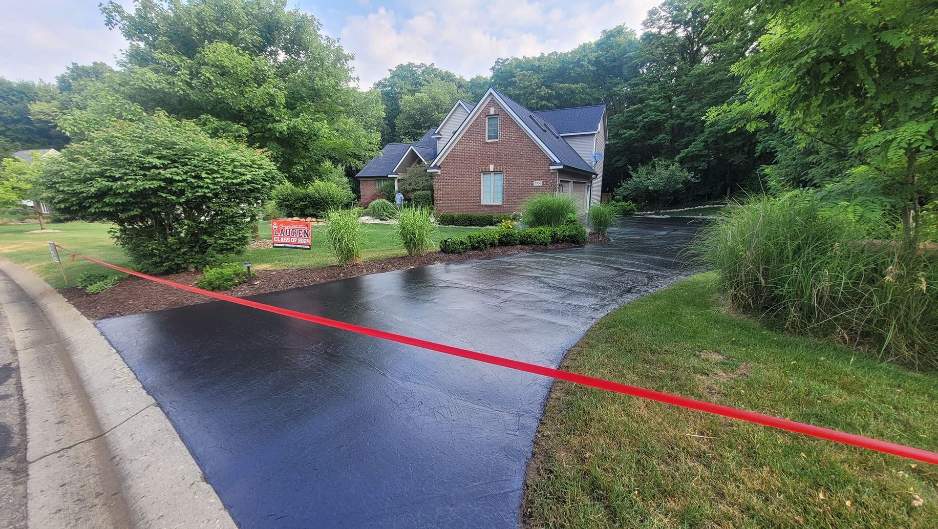 Driveway leading to a brick house with a dark roof, surrounded by green trees and shrubbery. Red caution tape across the driveway.