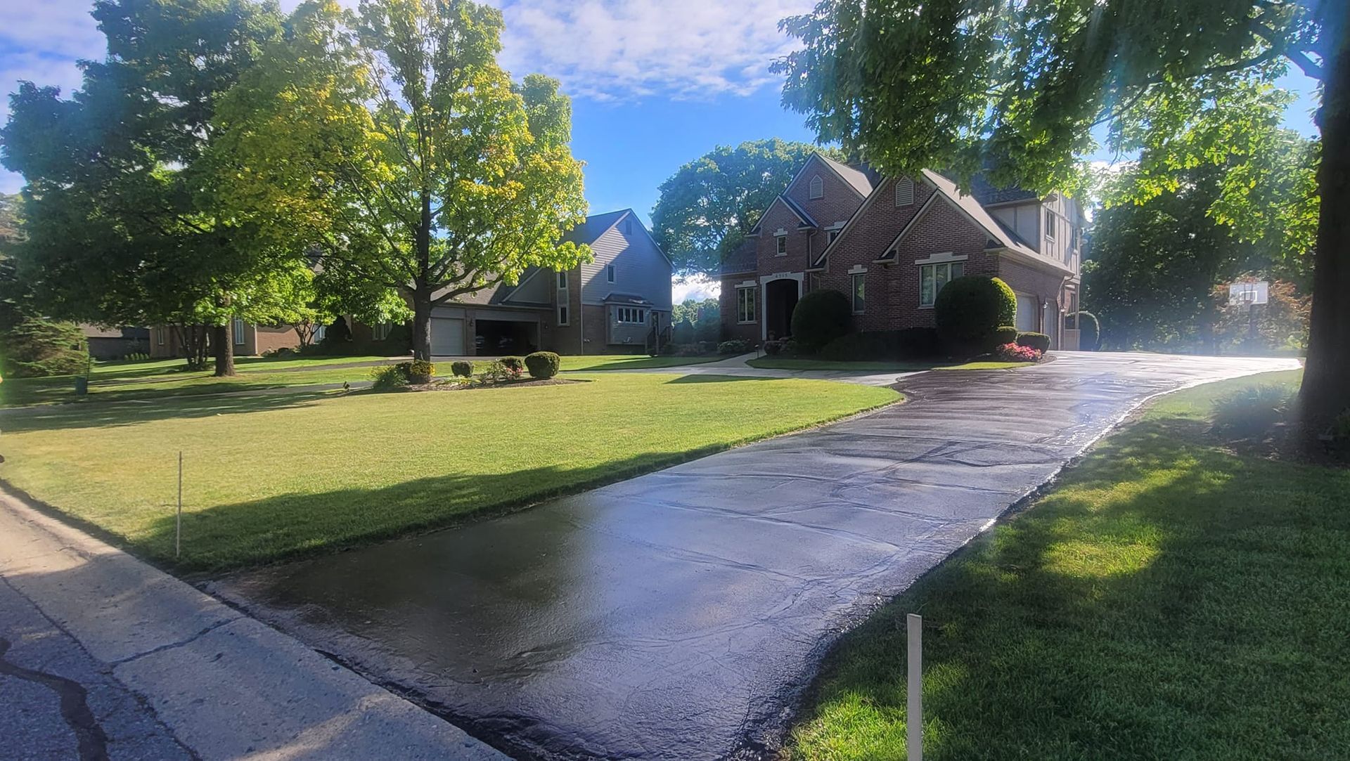 Residential street scene with wet asphalt driveway and green lawns under a bright blue sky, with two brick houses.