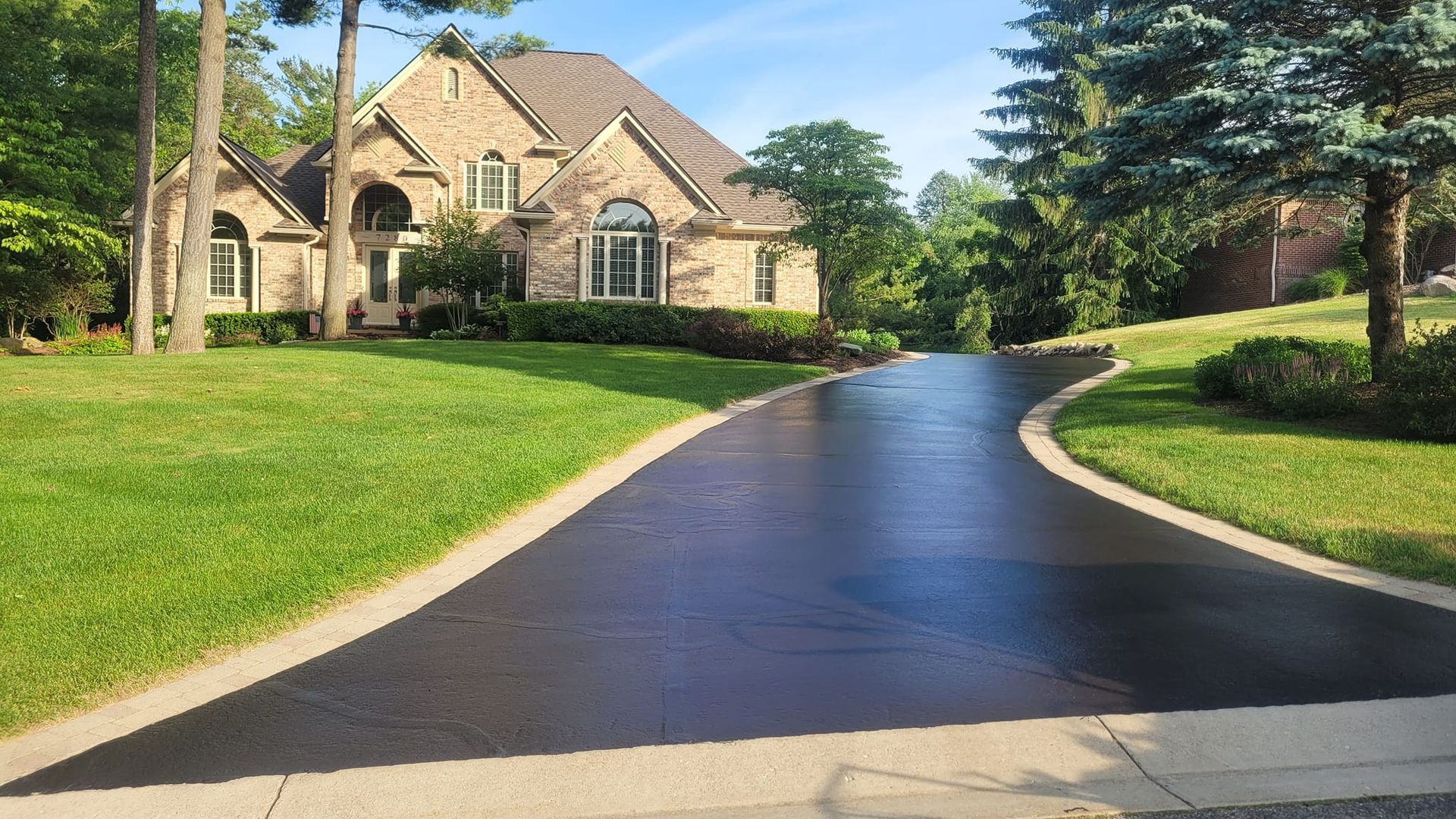 A brick house with a freshly sealed black driveway curves towards the front door, flanked by green lawns and trees.