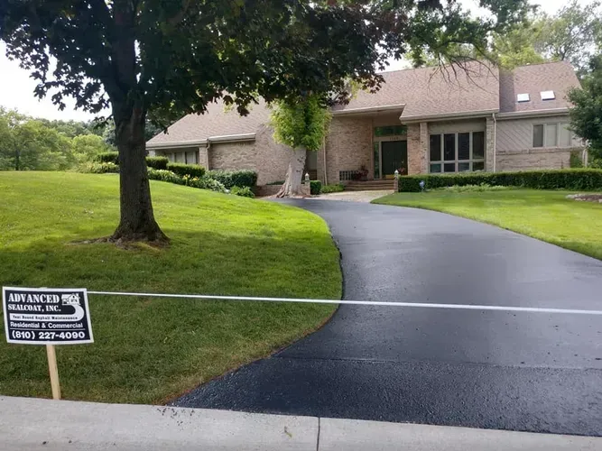 A house with a newly paved driveway, framed by green grass and a tree; a company sign is placed on the lawn.