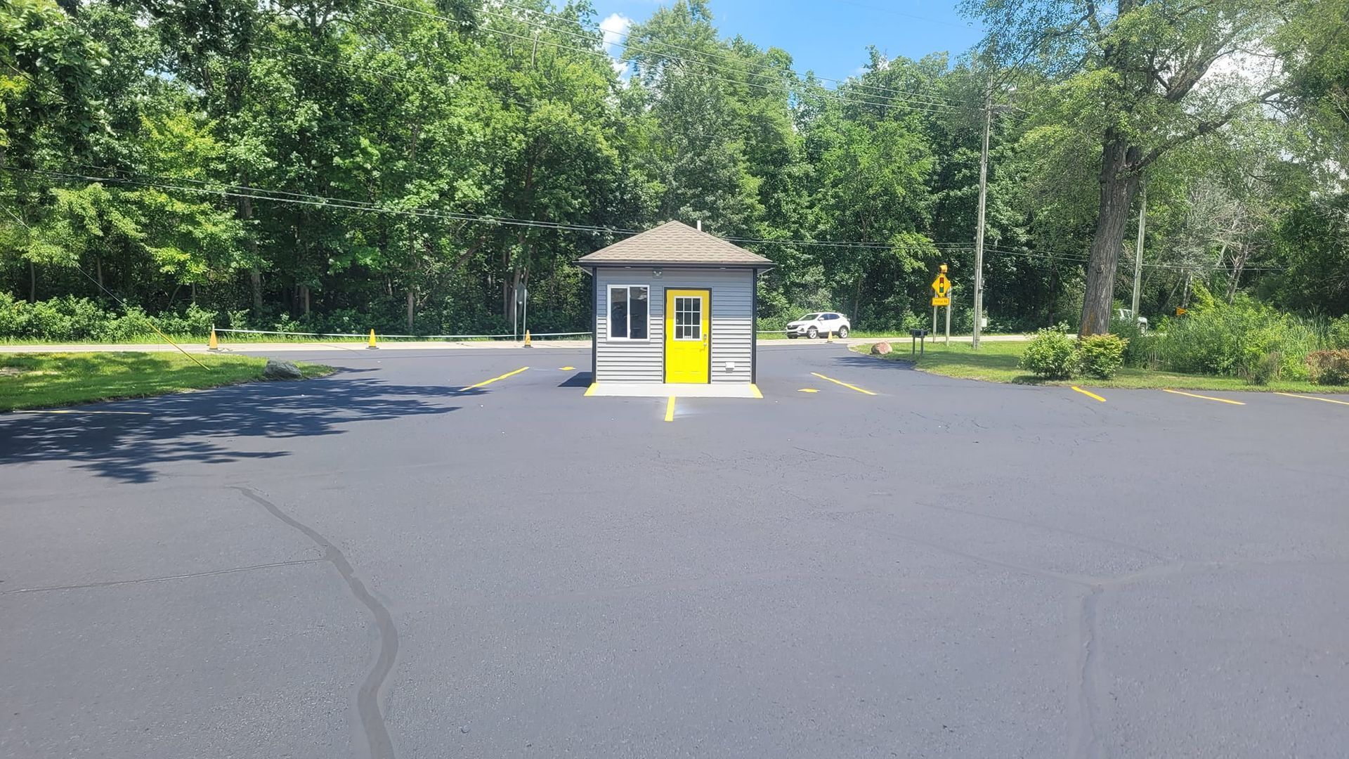 Small building with a bright yellow door and window in the middle of an empty parking lot. Trees line the background under a blue sky.