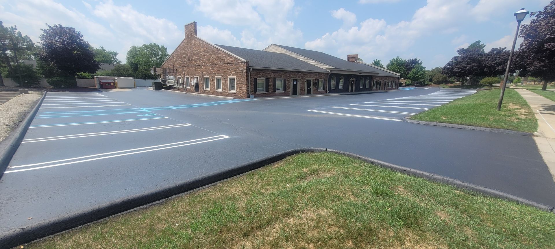 A newly paved parking lot with painted parking spaces, next to a building with a brick facade. Lush green grass surrounds the lot.