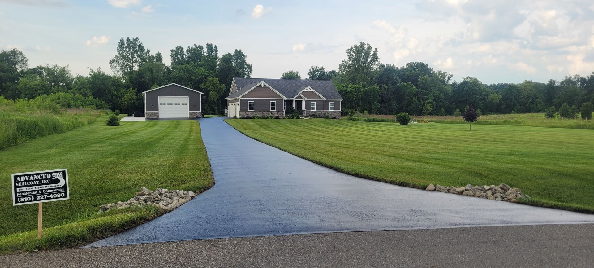A house with a long driveway and a garage. The house is surrounded by a green lawn and trees.
