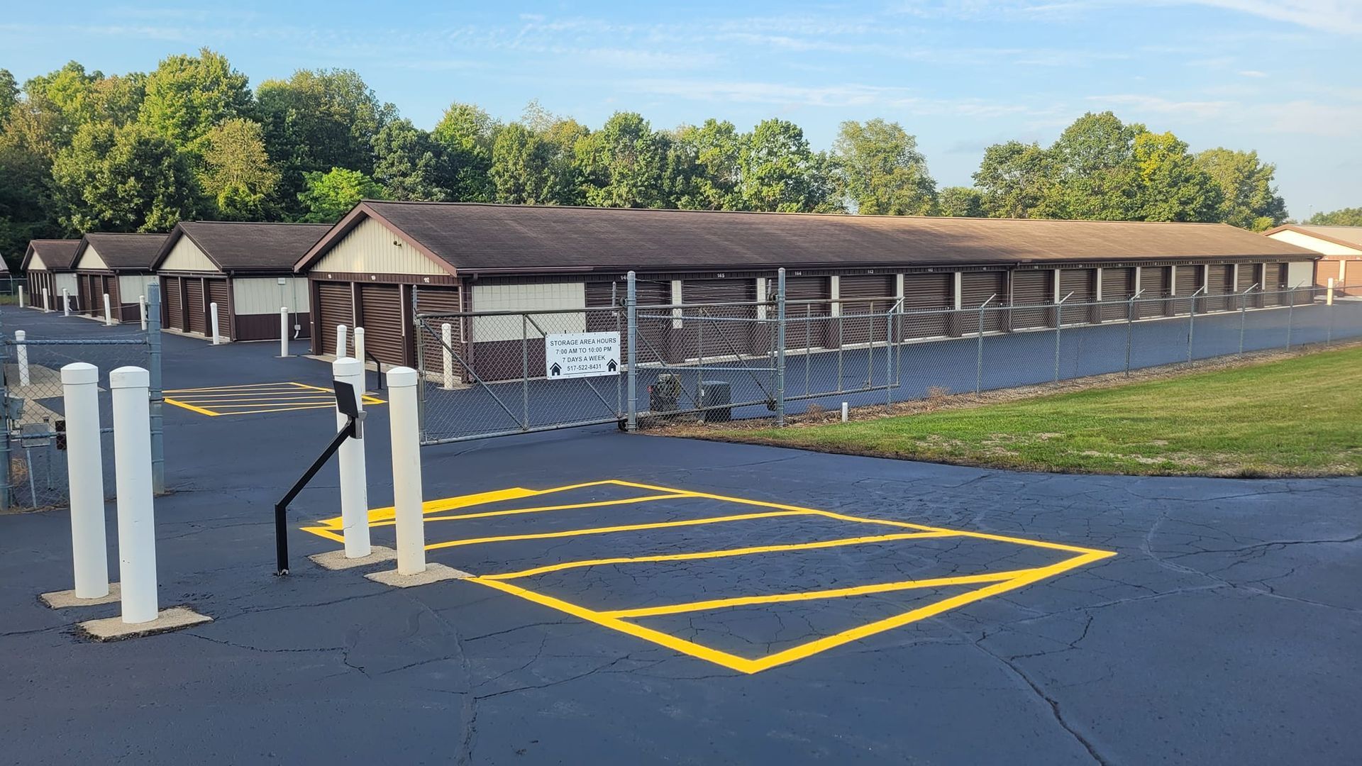Storage units with brown doors and roof, surrounded by a fence and parking area. Green trees and blue sky are in the background.