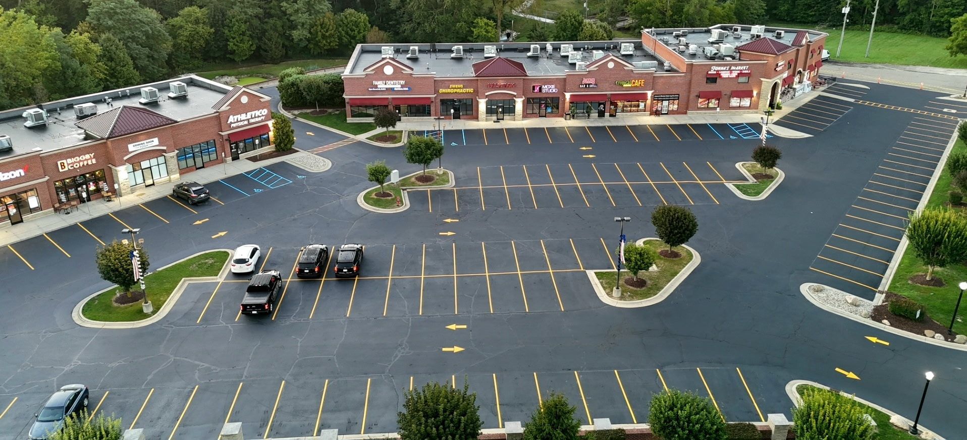 Aerial view of a parking lot with multiple stores. There are cars parked in the lot, and trees along the edges.