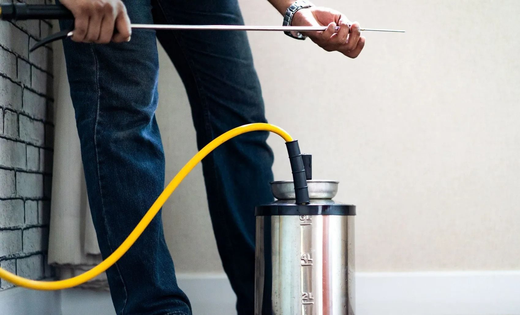 Person spraying insecticide with a tank sprayer near a wall.