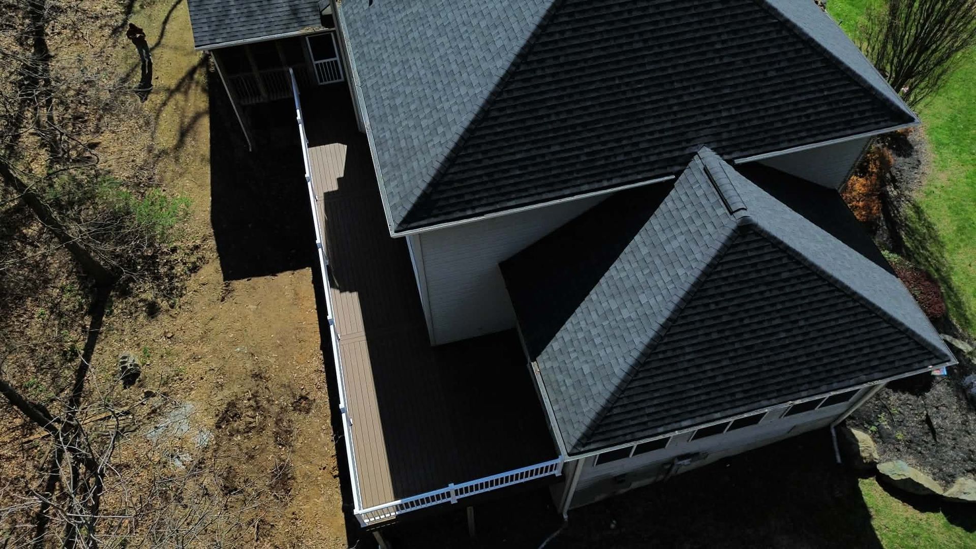 An aerial view of a house with a dark gray shingled roof, a large attached deck, and a surrounding wooded landscape.