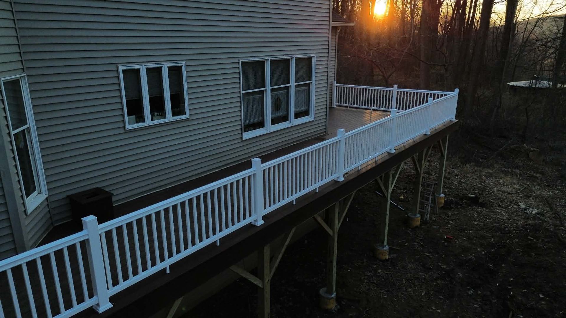 A raised wooden deck with white railings attached to the side of a light-colored house during sunset.