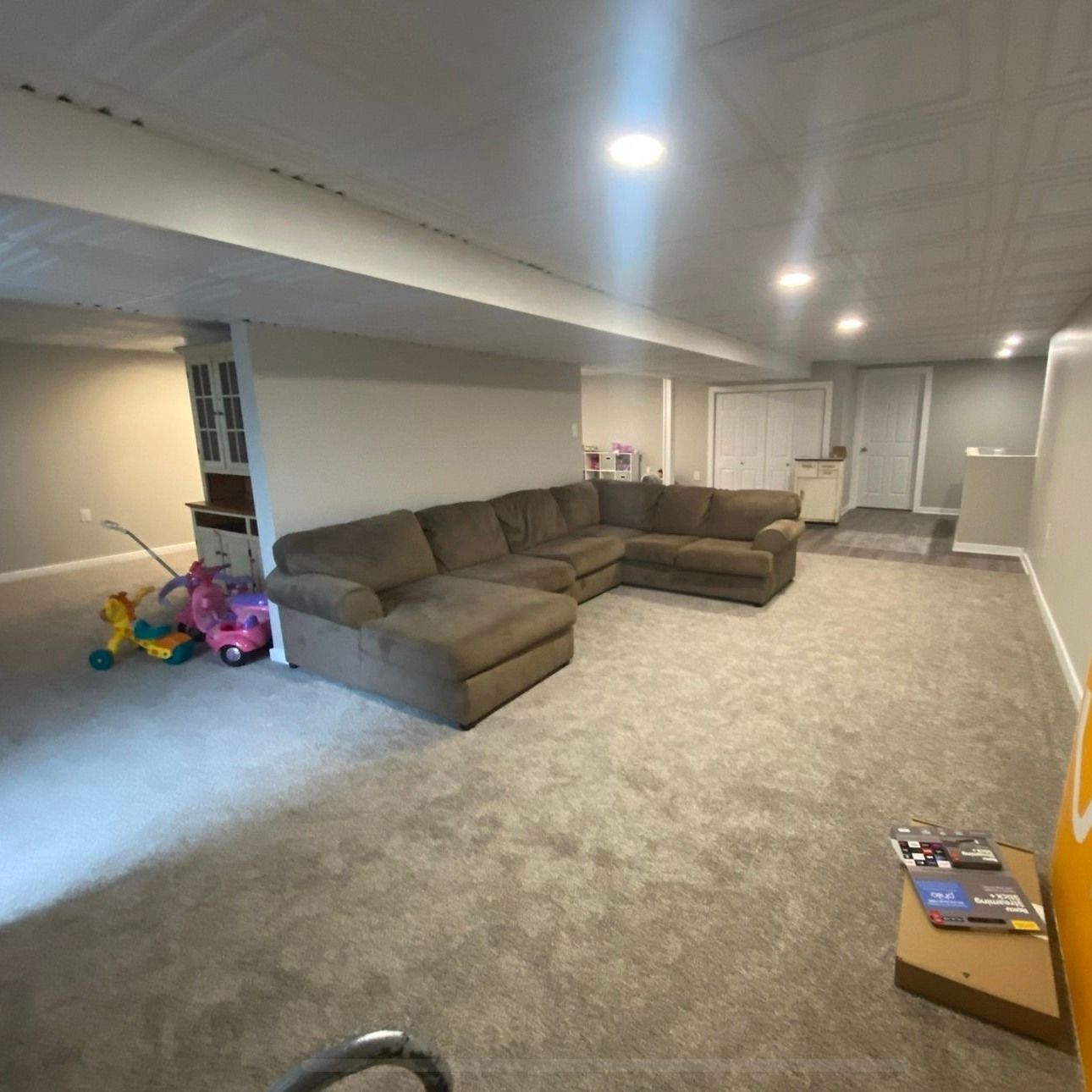 A living room in a basement with a sectional couch and a pink toy bike.