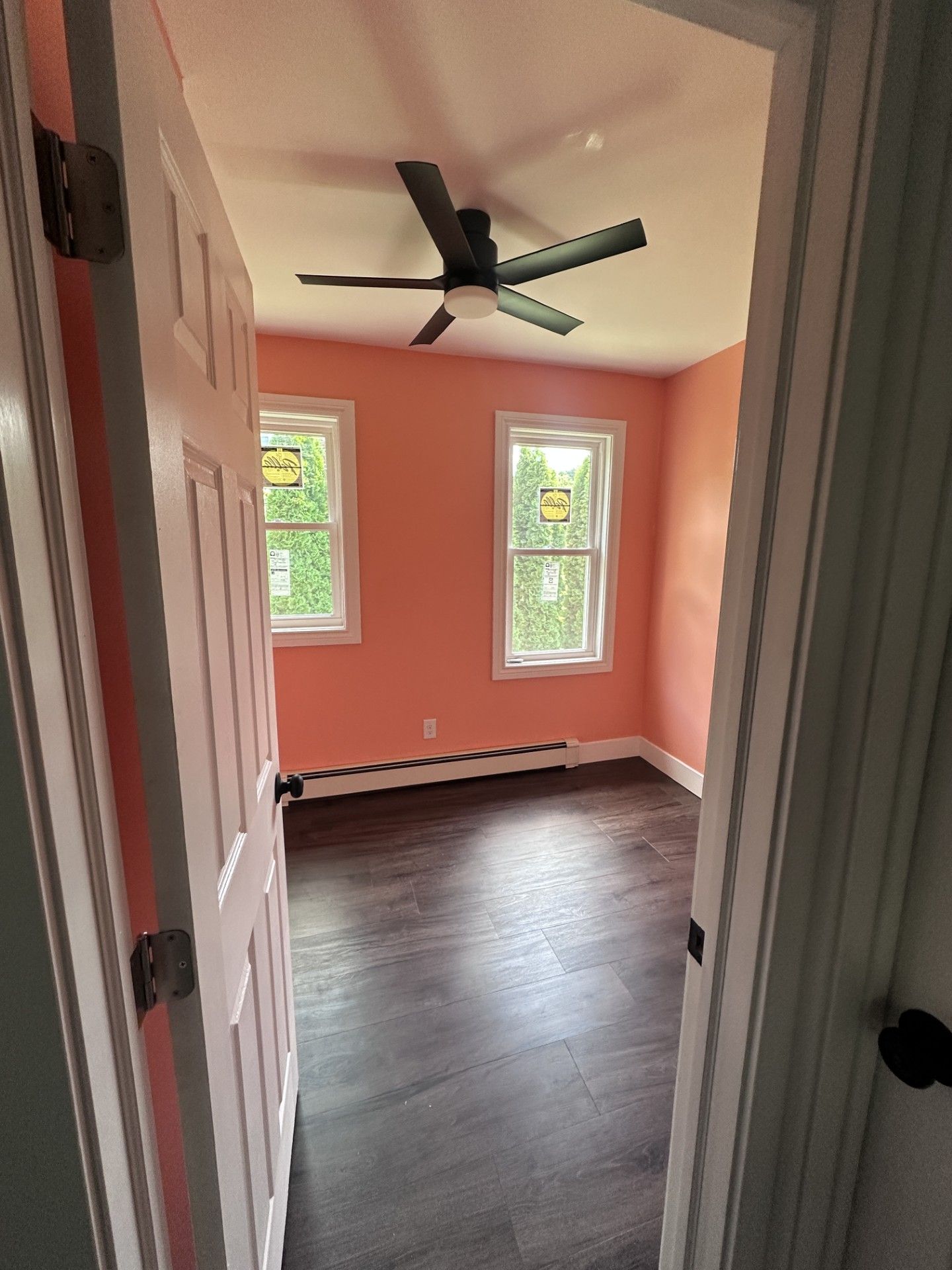 A view through an open doorway into an empty bedroom with salmon-colored walls, dark wood floors, and a ceiling fan.