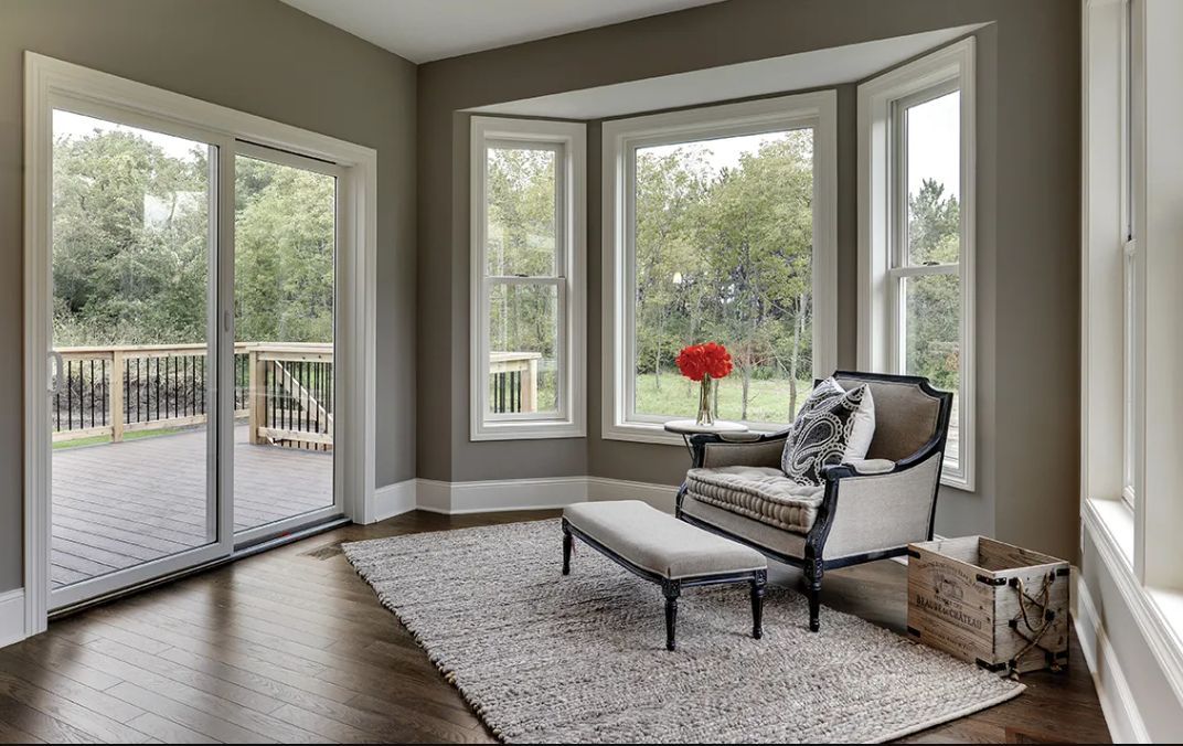 A sunlit room with hardwood floors, a bay window, a glass door to a deck, and an armchair with a footstool on a rug.