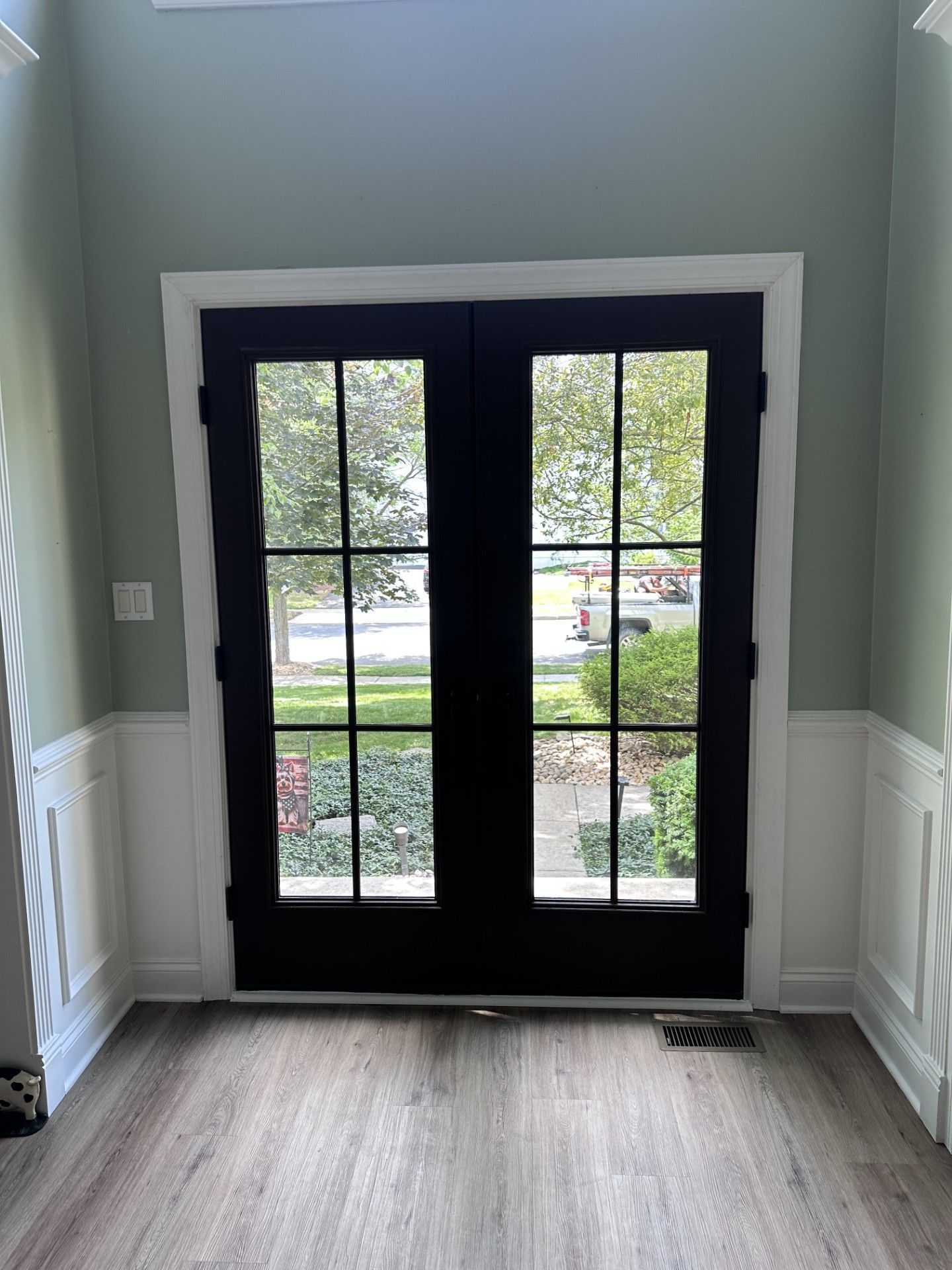 A view from inside a home looking out through black French double doors at a front yard with trees and a parked vehicle.
