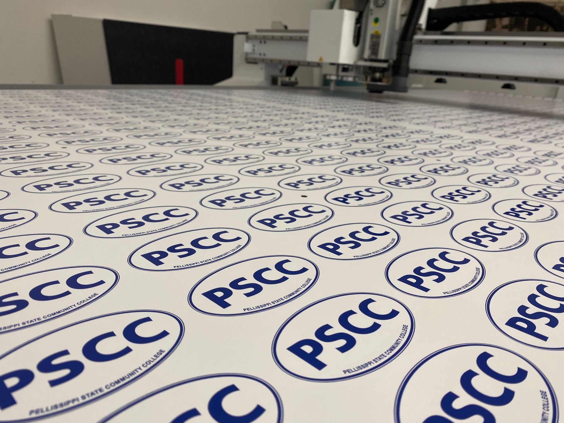 A machine cutting out blue and white PSCC logo stickers from a printed sheet.