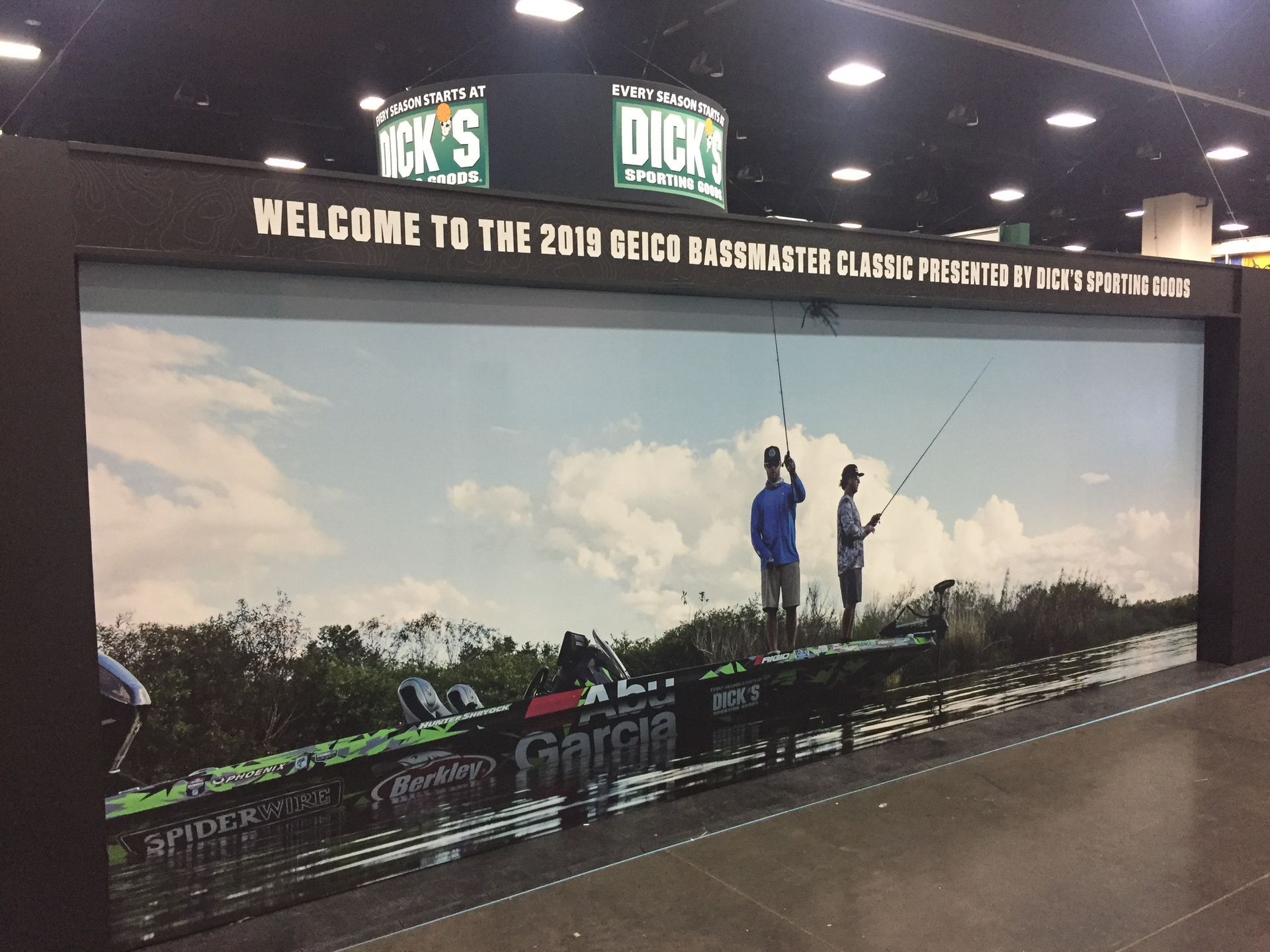 DICK'S Sporting Goods display at 2019 GEICO Bassmaster Classic. Two people fishing on a boat, cloudy sky background.