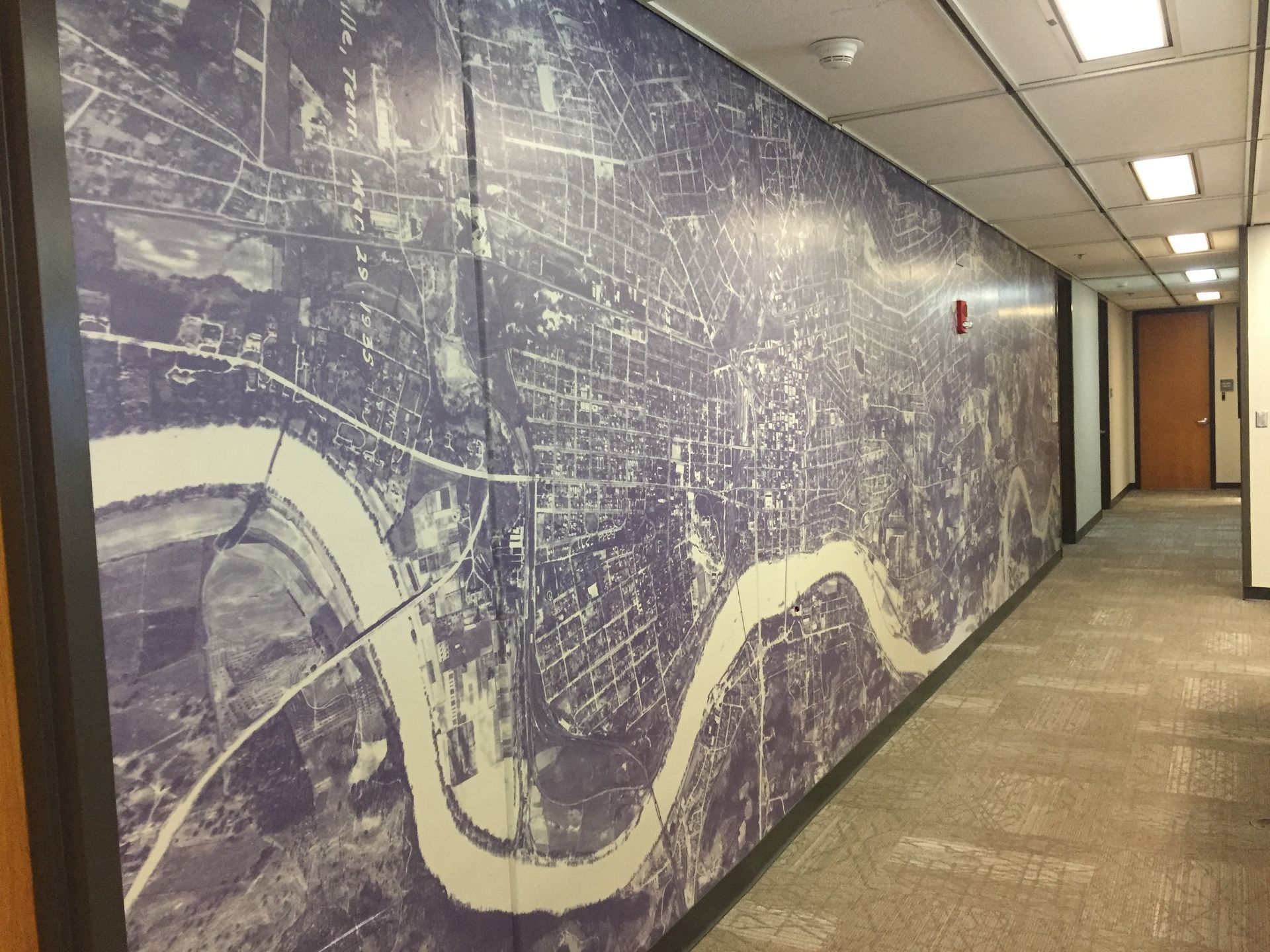 Hallway with a large wall mural of a city map; river visible. Fluorescent lights and a brown door.
