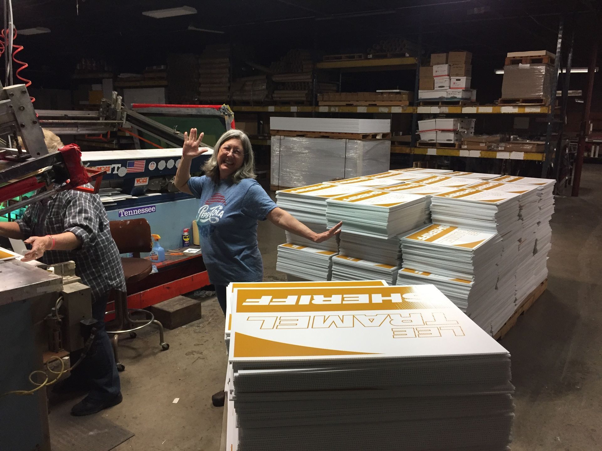 Woman waves near a stack of printed signs in a warehouse. Machinery and shelves of materials are visible.