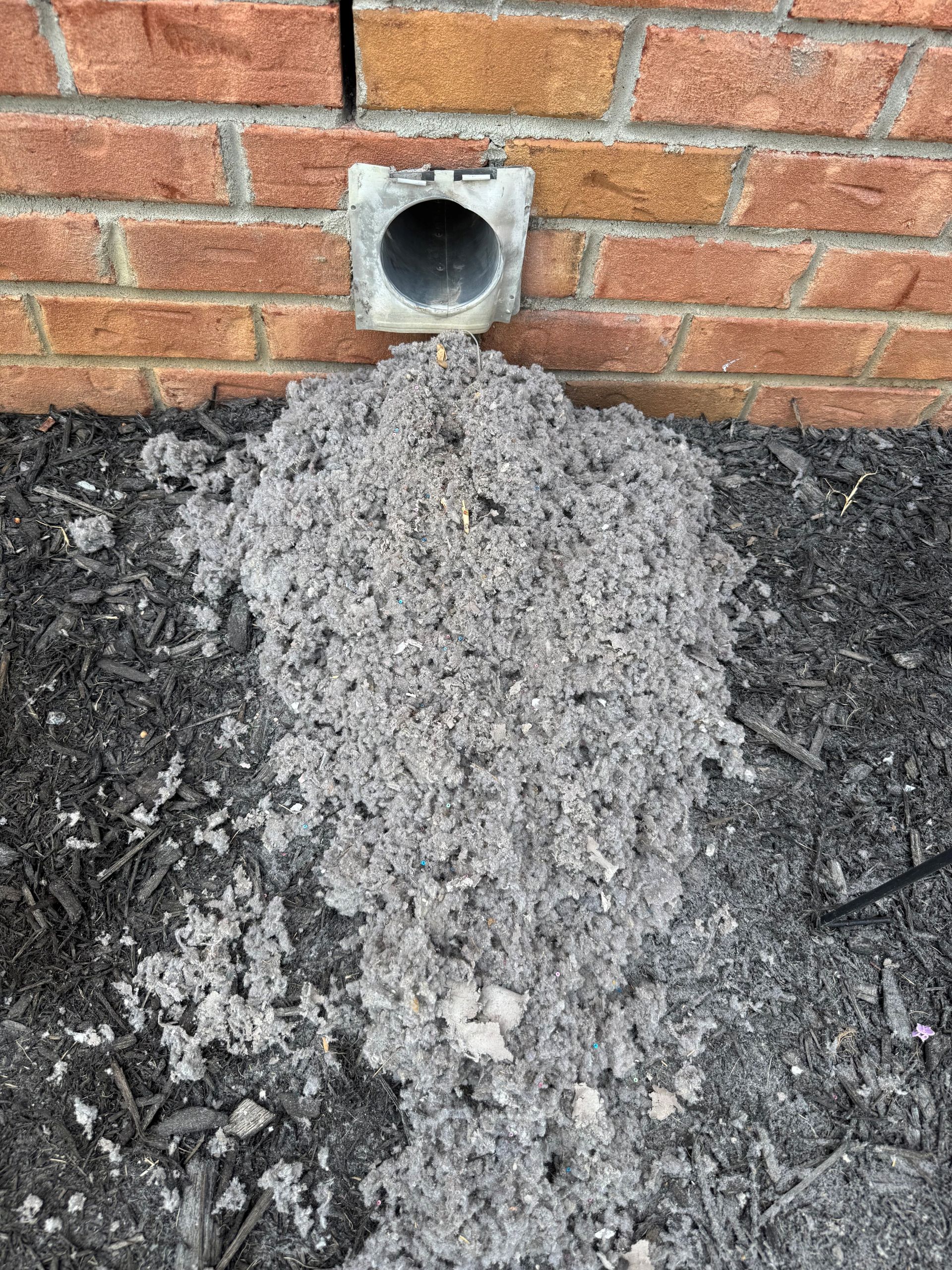 Grey lint pile spilling from dryer vent on a brick building's exterior.