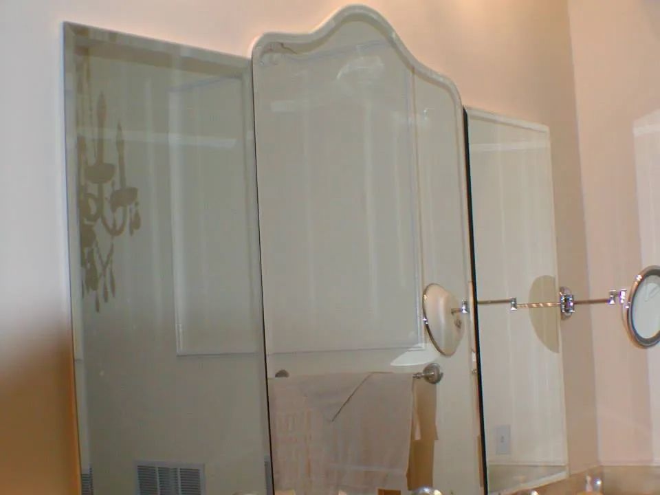 Three-paneled bathroom mirror; the center mirror is ornate. Reflects a chandelier and a towel bar.