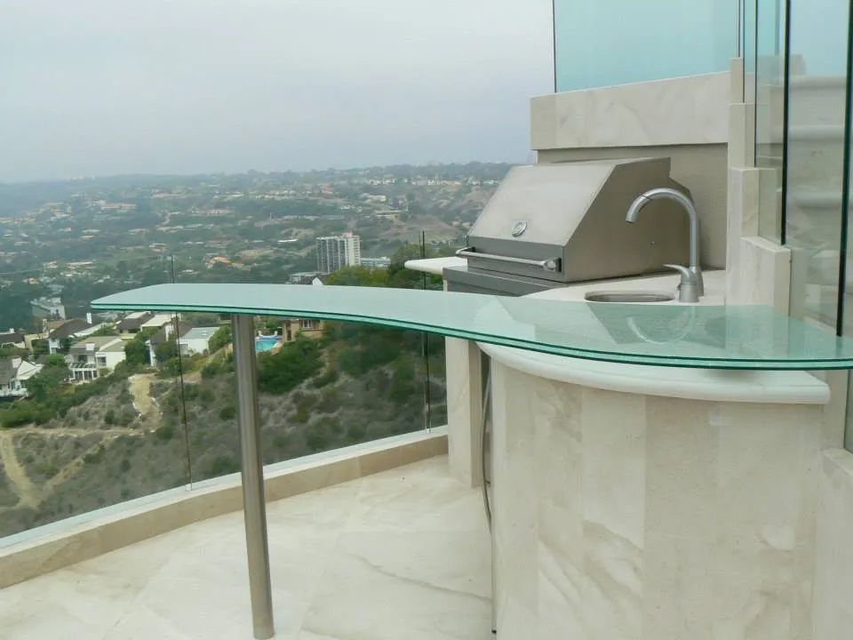 Outdoor kitchen with a grill, sink, and glass countertop overlooking a city.