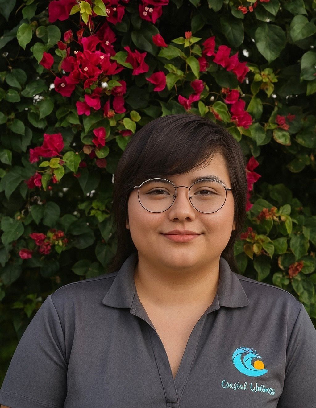 Woman with glasses, short dark hair, smiling, in front of red flowers and green leaves, wearing a grey polo shirt with a wave logo.