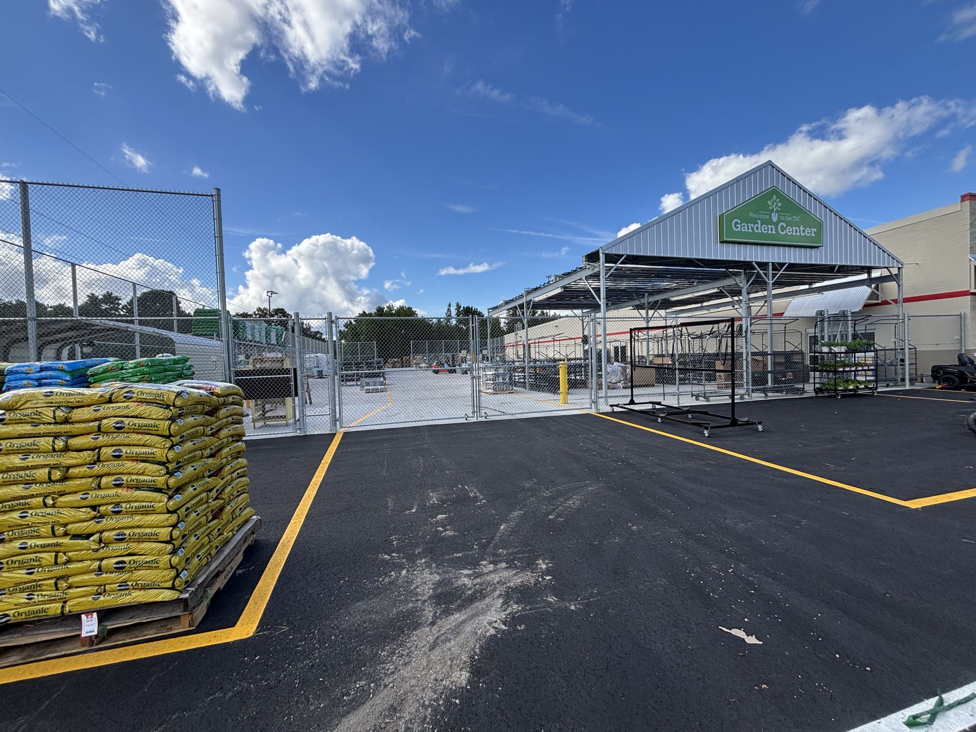 Exterior view of a garden center with a metal fence, empty displays, and stacks of yellow bags.