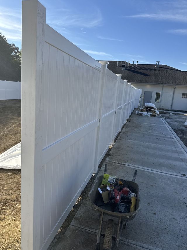 White vinyl fence being built alongside a concrete walkway; a wheelbarrow with tools sits nearby.