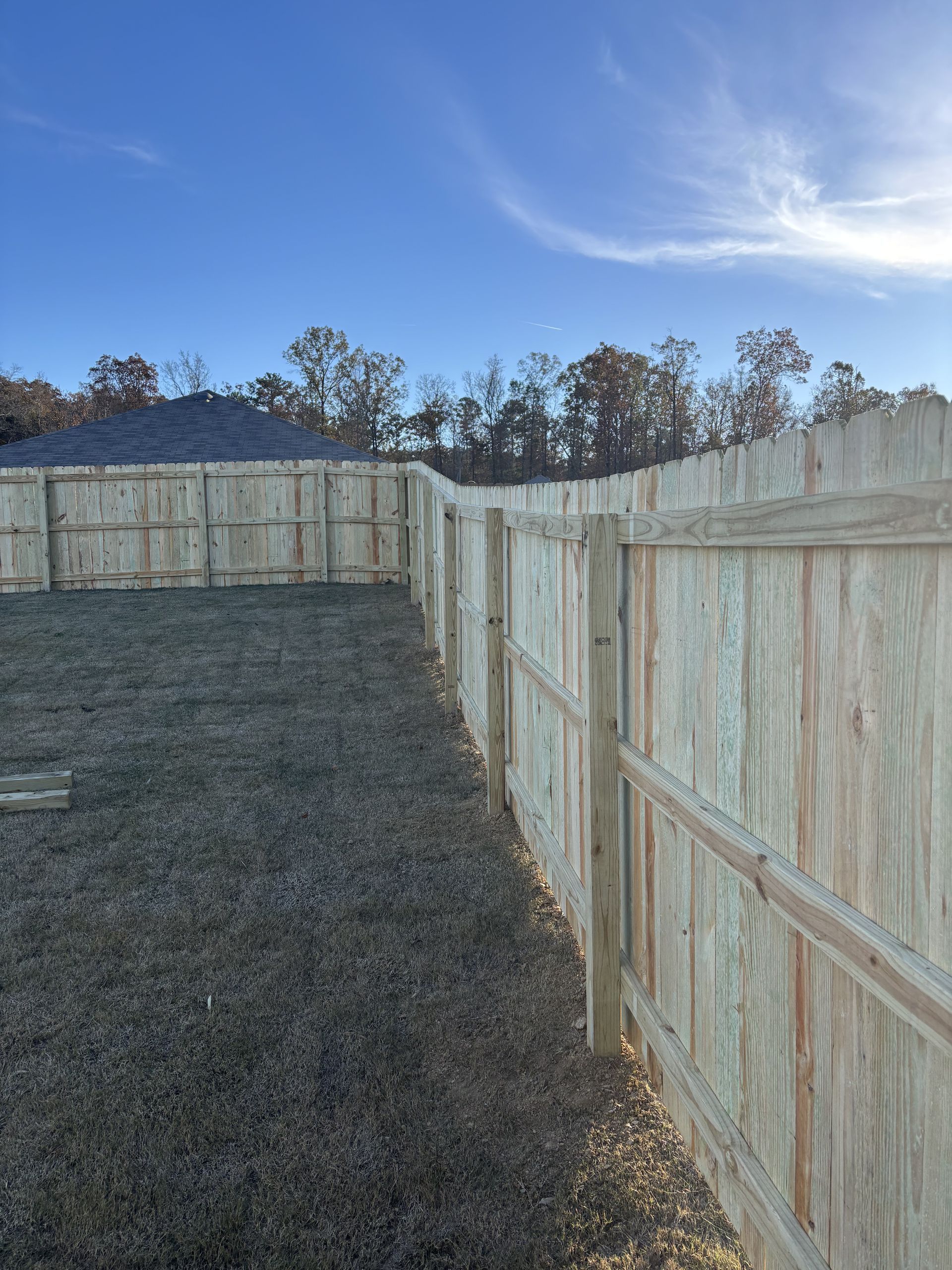 Wooden fence surrounds a yard with brown grass on a sunny day. Trees and blue sky in the background.