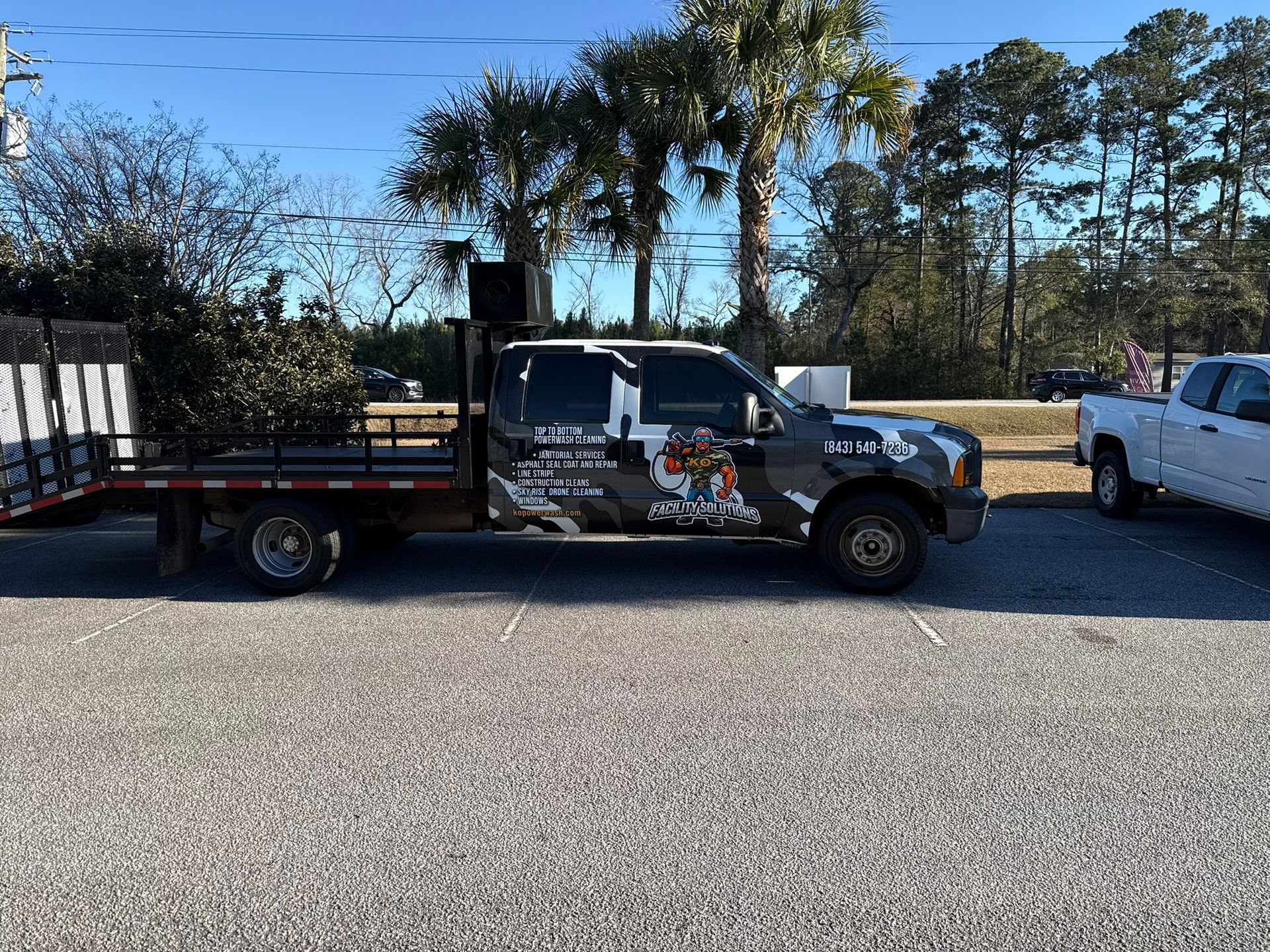 Black flatbed tow truck parked on gravel beside a white pickup under a clear blue sky.