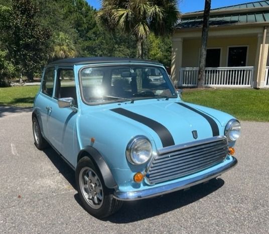 Light blue classic Mini Cooper with black racing stripes, parked in front of a building on a sunny day.