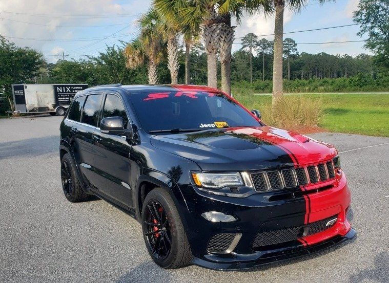 Black and red Jeep Grand Cherokee SRT SUV parked on a paved area, with palm trees and a trailer in the background.