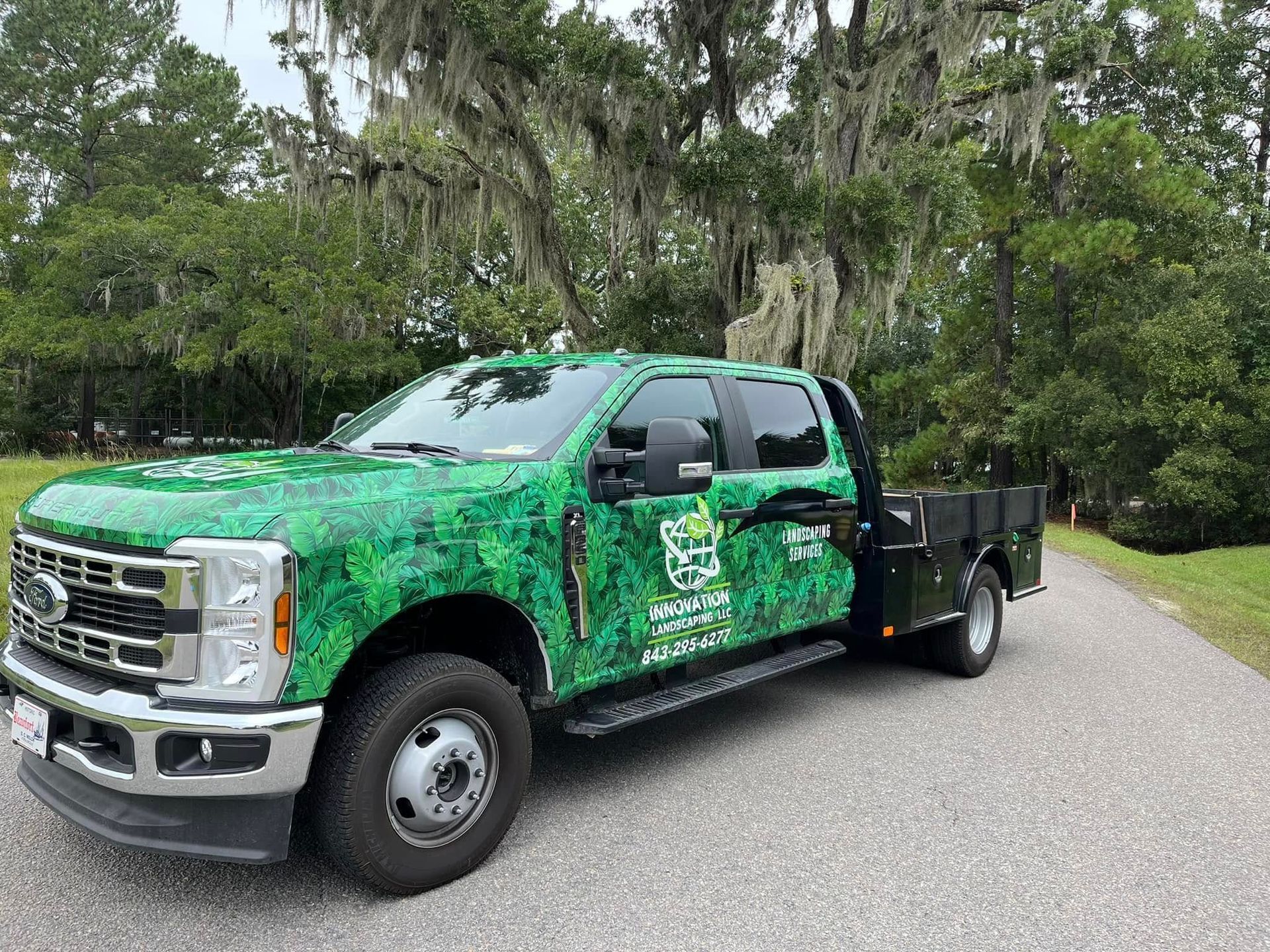 Green-wrapped work truck with landscaping business logo parked on a paved road, trees in the background.