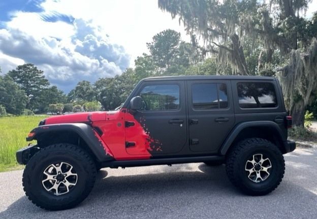 A red and black Jeep Wrangler with large tires parked on a road, under a cloudy sky.