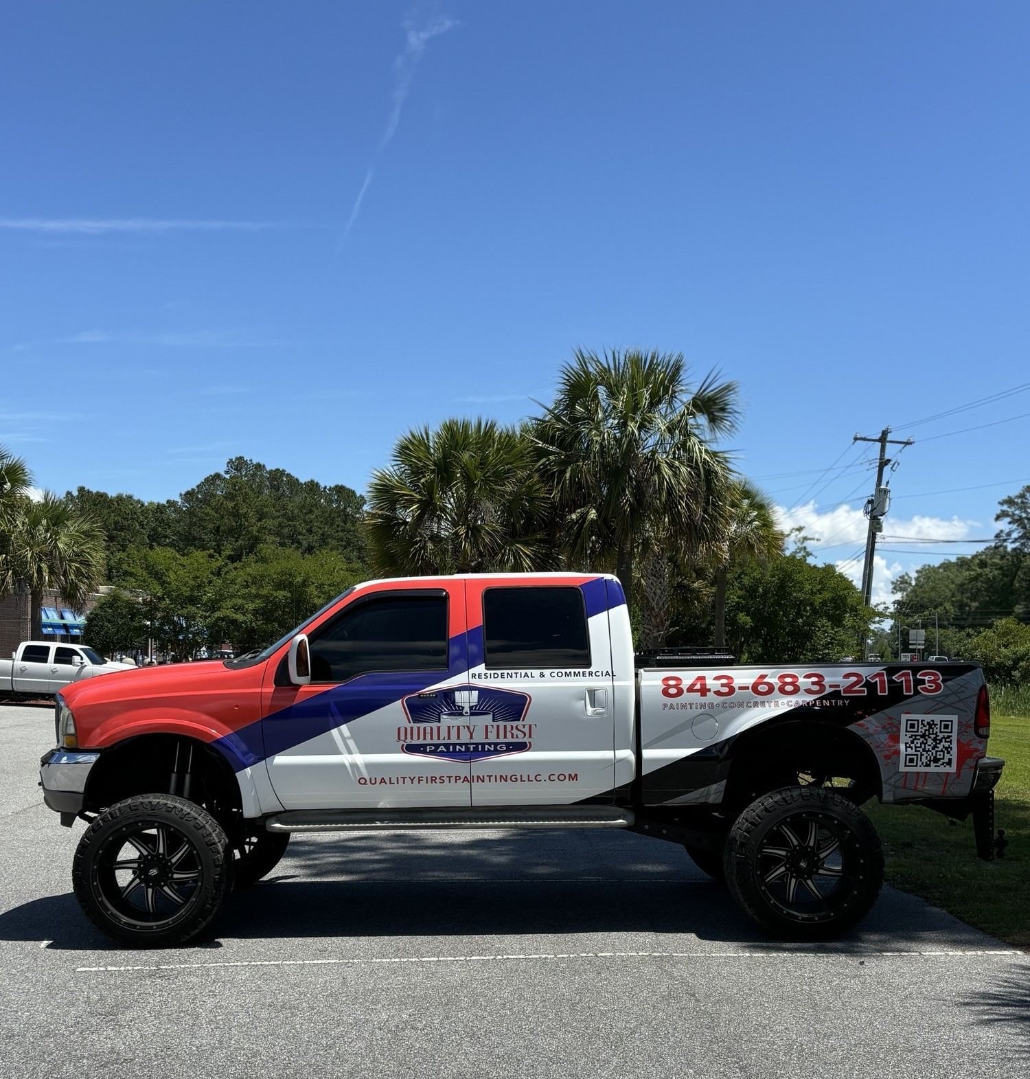 Red, white, and blue custom pickup truck with company logo and phone number parked in front of trees under a blue sky.